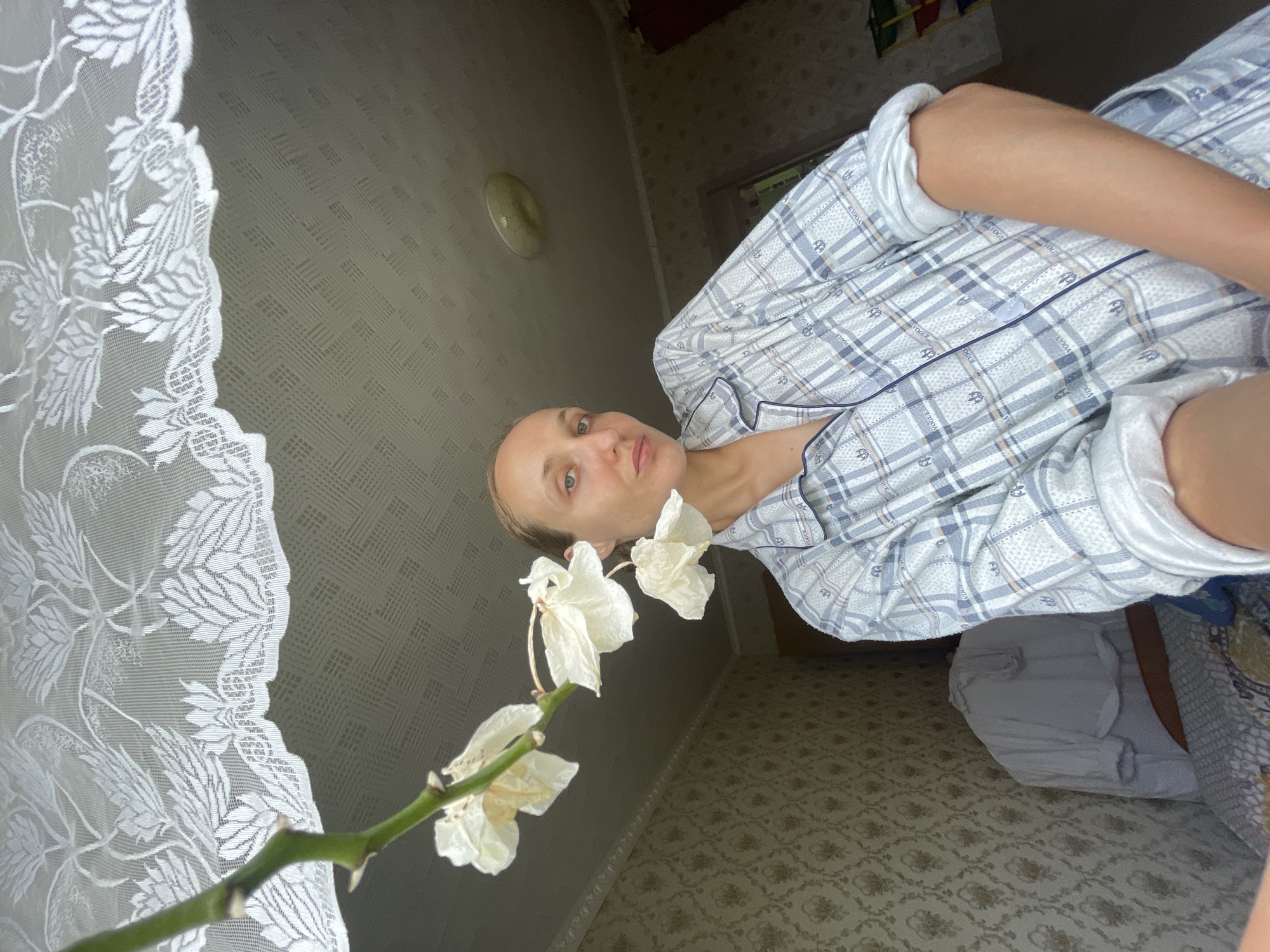 A woman, appearing to be in her late twenties or early thirties, with fair skin and light brown hair pulled back from her face, looks directly at the camera. She is wearing blue and white plaid pajamas with a navy trim. She is indoors, positioned in front of a window covered by a white lace curtain. A dried white orchid plant hangs from the upper left of the frame, in front of the curtain. The room appears to be a bedroom, with patterned wallpaper visible on the left wall and a light fixture on the ceiling. A bed with patterned bedding is partially visible in the lower left. The overall lighting suggests it is daytime. The location is identified as Sharkan, Russia.