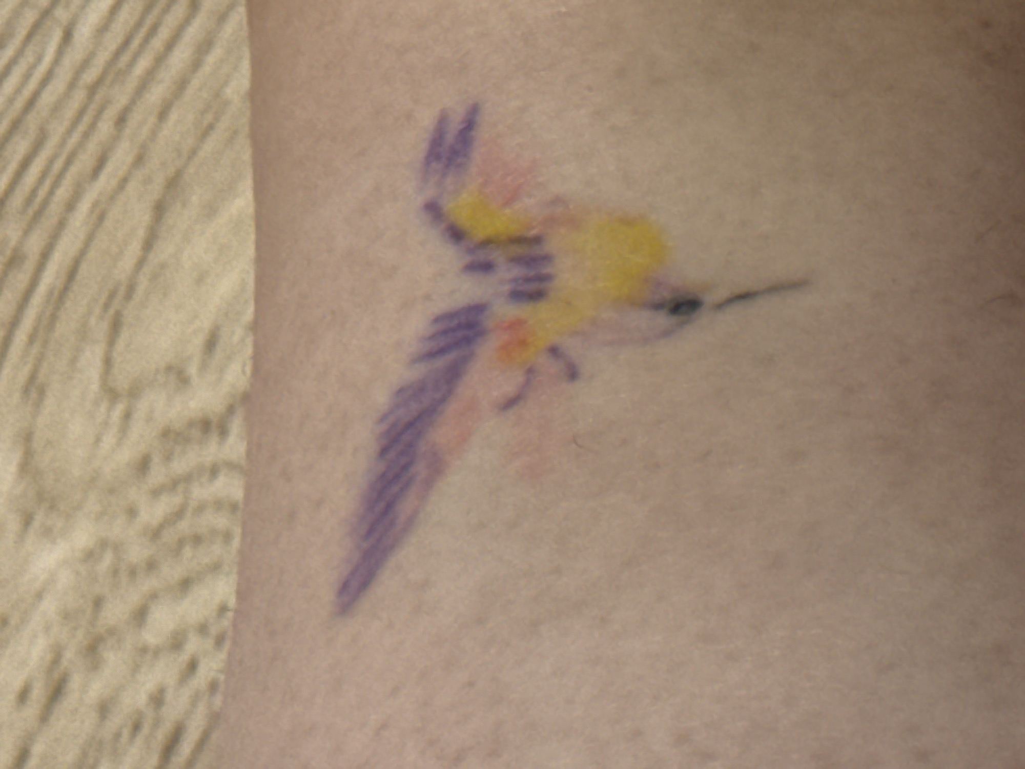 The image is a close-up of a tattoo on human skin. The tattoo depicts a hummingbird in flight, rendered with purple, yellow, and pinkish-orange colors. The hummingbird is shown with its wings spread and its beak pointed forward. The background is the texture of the skin, with a slight shadow or texture pattern visible on the left side of the frame, possibly from a piece of fabric or a textured surface. There is no discernible text in the image. The focus is entirely on the tattoo and the skin it is on. The lighting appears to be from a single source, casting subtle shadows and highlighting the texture of the skin and the ink. The overall impression is a small, artistic tattoo with a vibrant color palette.