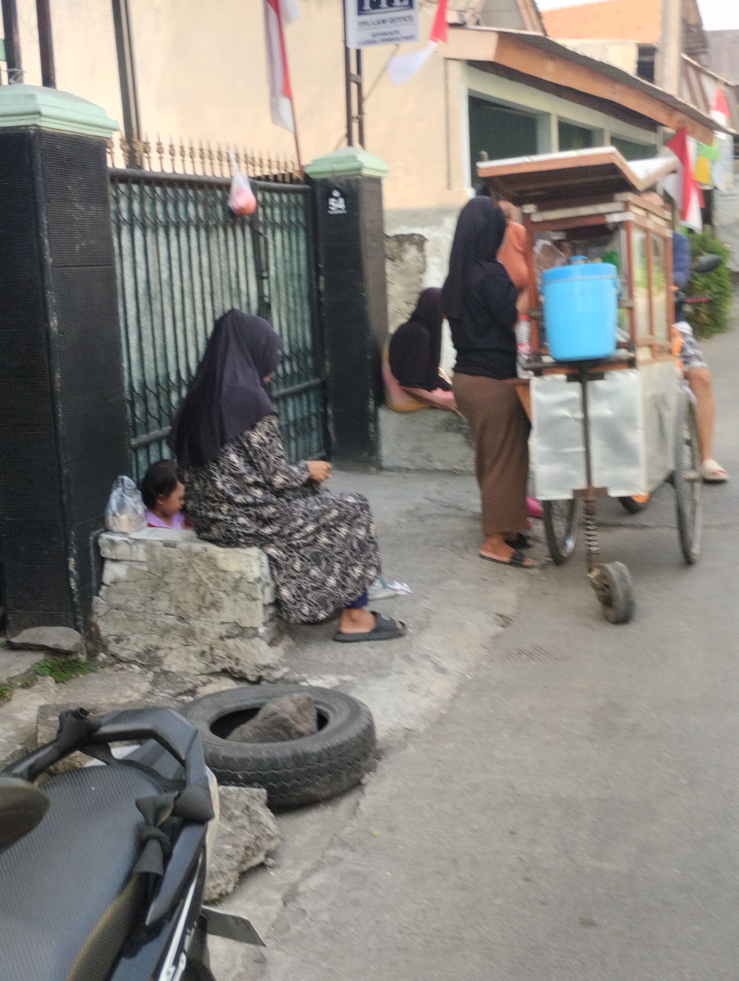 The image depicts an outdoor scene likely in Bekasi, Indonesia, on a street during daylight. In the foreground, a portion of a black motorcycle is visible on the left. A large, discarded tire lies on the ground to the right of the motorcycle, with a rock inside it.
Further into the scene, a woman wearing a dark hijab and a patterned dress is seated on a concrete ledge on the left side of the image. Beside her, a small child in a pink top is seated. A translucent plastic bag hangs from a black metal gate in the background.
To the right of the seated woman, a food cart with a blue container and a wooden top is visible. A person wearing a black hijab and brown pants is standing next to the food cart. Another person, with their legs visible, is standing behind the food cart. The environment appears to be a street with a building in the background, adorned with Indonesian flags. The ground is paved with asphalt and concrete.