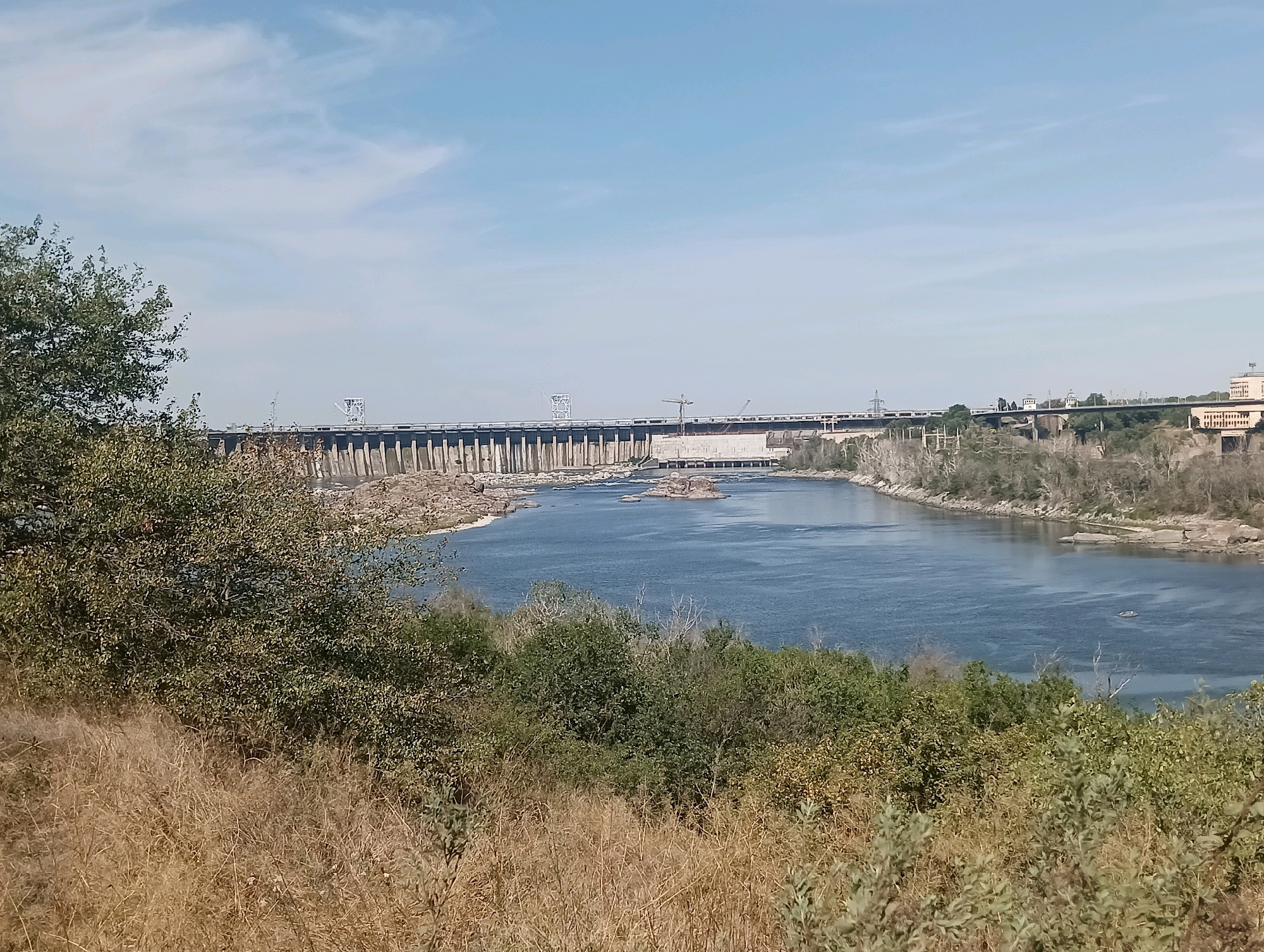 This is a wide-angle, eye-level, outdoor, daytime photograph of a large hydroelectric dam and the body of water it impounds. The dam is a long, grey, concrete structure that stretches across the frame. It has a tall, imposing appearance with numerous pilings and visible structures on its surface. Behind the dam, a bridge or elevated road is visible, supported by columns.
The water in front of the dam is a deep blue, reflecting the sky. There are several rocky islands scattered in the water. The foreground of the image is dominated by dry, golden grass and various green bushes and trees. The sky is a light blue with some wispy white clouds.
The scene appears to be a landscape shot, likely taken from a vantage point on a hill overlooking the dam and river. The overall impression is one of industrial scale meeting natural landscape. No people are visible in the frame.