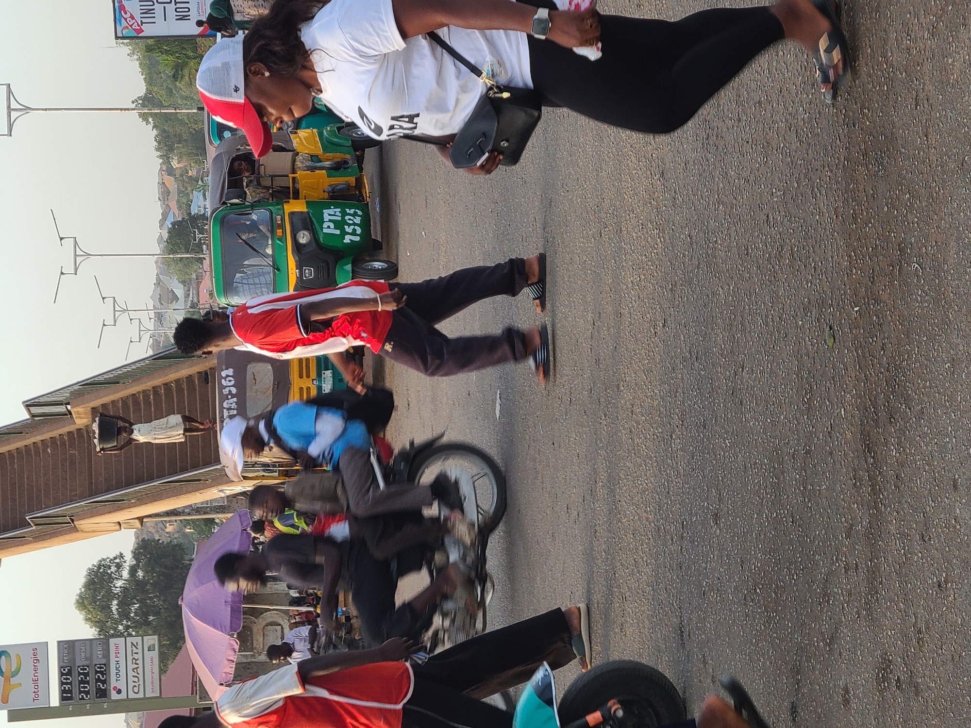 This media file captures a vibrant street scene in Jos, Nigeria, likely during the golden hour of either early morning or late afternoon, characterized by the warm, soft lighting.
The primary subjects are numerous people and vehicles engaged in daily activities along a paved road. In the foreground, a woman wearing a white t-shirt, black leggings, and a red and white baseball cap is walking, carrying a small bag. Behind her, a man in a red and white shirt also walks on the roadside. The scene is bustling with movement, as indicated by the slight motion blur on people and vehicle wheels.
Several modes of transport are visible: multiple tricycles, commonly known as "keke Napep" in Nigeria, and motorcycles. One prominent light-colored tricycle bears the text "PTA 7525" on its front. Another green and yellow tricycle is parked or moving slowly. In the mid-ground, a man on a motorcycle is clearly seen, and further back, another individual carries an item balanced on their head, a common sight in the region. A purple umbrella suggests the presence of a vendor or shade for individuals.
Notable details include a "Total Energies" fuel station sign on the left side of the image, displaying fuel prices: "1309 PETROL," "2020 DIESEL," and "22.0 KERO." Other text on the sign includes "TOUCH POINT" and "QUARTZ." In the background, a billboard with partial text "AP" and "TINU" is visible, along with building structures. The general setting is an urban environment with buildings and distant hills under a clear sky. The overall impression is one of everyday life and commute in a busy Nigerian city.