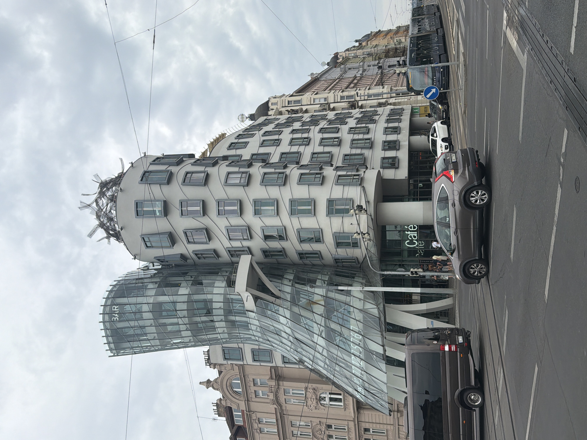The image captures the Dancing House, a unique modern architectural landmark located in Prague, Czechia. The building, characterized by its unconventional, fluid design resembling two dancers, stands prominently on the right side of the frame. Its glass facade on the left tower curves outwards, revealing multiple levels. To the right, the building features a series of repetitive, dark-framed windows. The sky above is overcast with clouds. In the foreground, a street scene unfolds. A brown van is visible on the left, and a grey SUV is in the center, both driving on the road. Further down the road, a modern tram is depicted. Several pedestrians are seen near the entrance of the Dancing House, some appearing to be entering or exiting the building, where a sign for "Café" is visible. The street is paved with asphalt and marked with white lines. Other traditional European buildings line the street in the background.