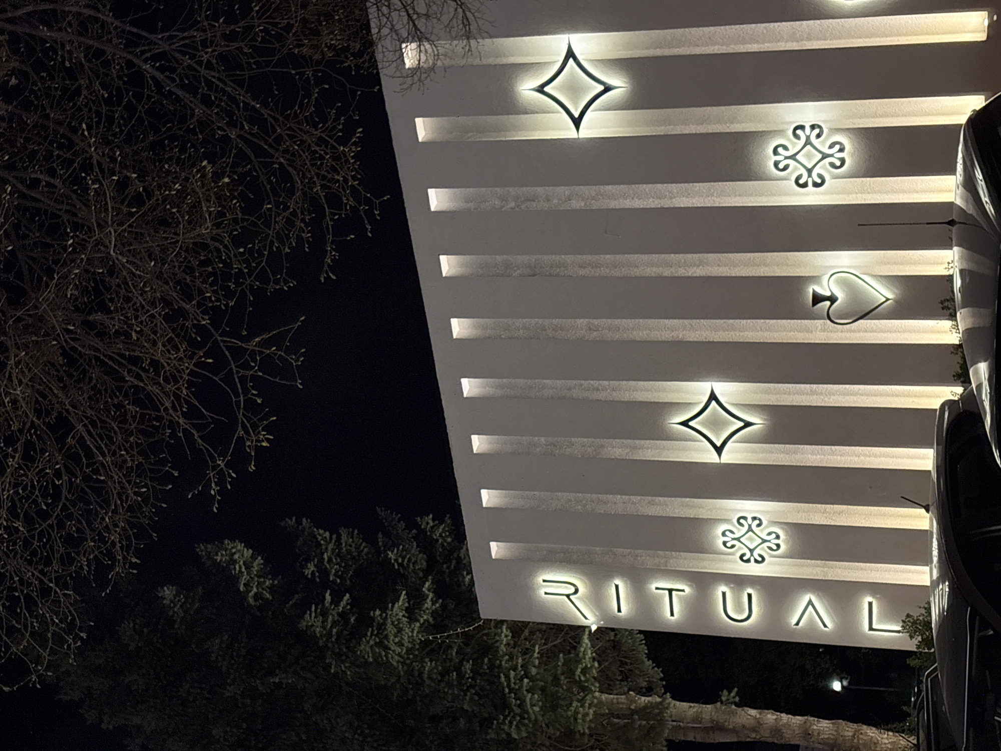 A nighttime shot shows a portion of a building with the word "RITUAL" illuminated at the bottom. Above the name, there are four distinct symbols, each also illuminated: a diamond shape, a floral-like design, a spade, and another diamond shape. The building is white, with horizontal lines that create a distinct layered effect. A bare tree is visible on the left side of the frame, and some greenery is at the bottom, partially obscuring the base of the building. A dark, reflective object, potentially a vehicle, is positioned on the right edge of the frame, adding depth to the scene. The location is Sofia, Bulgaria.