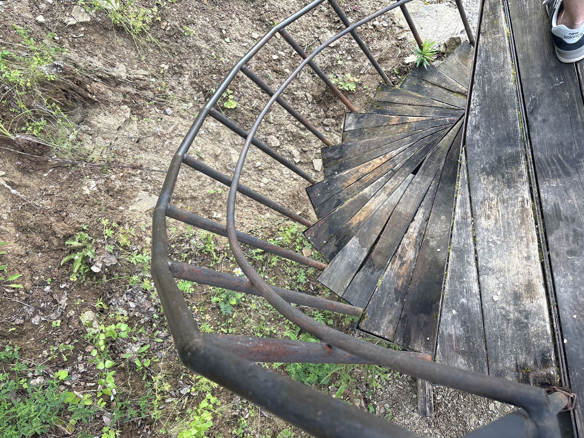 An outdoor, top-down shot shows a rustic, spiral staircase constructed of weathered wooden planks and a dark metal railing. The staircase is set against a backdrop of dry, rocky earth and sparse green vegetation. The wooden treads are worn and uneven, with some appearing damp and mossy. The metal railing is also aged, showing signs of rust. The surrounding ground is covered in dirt, small stones, and patches of grass and small weeds. In the lower right corner, the edge of a person's foot wearing a New Balance sneaker is visible on one of the wooden planks, indicating someone is standing on or near the staircase. The scene evokes a sense of disuse and natural integration, suggesting the staircase may be part of an older structure or an access point to a natural area. There is no specific information provided within the image to pinpoint its exact location within the city of Lyady, Russia.