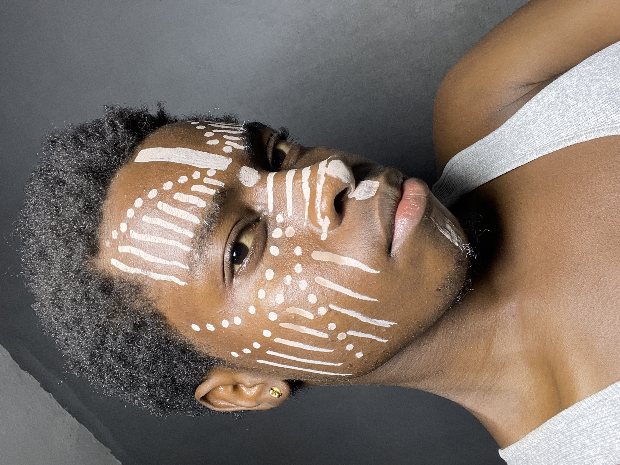 The image is a close-up, portrait-style shot of a person with dark skin and short, curly hair. The person is wearing a white tank top. Their face is adorned with white tribal-like markings consisting of short horizontal lines and dots. They have a gold stud earring in their left ear. The background appears to be a dark gray or black color. There is no indication of the location being Nkpor, Nigeria. The image primarily focuses on the person's face and upper body, with the white markings being a prominent feature.