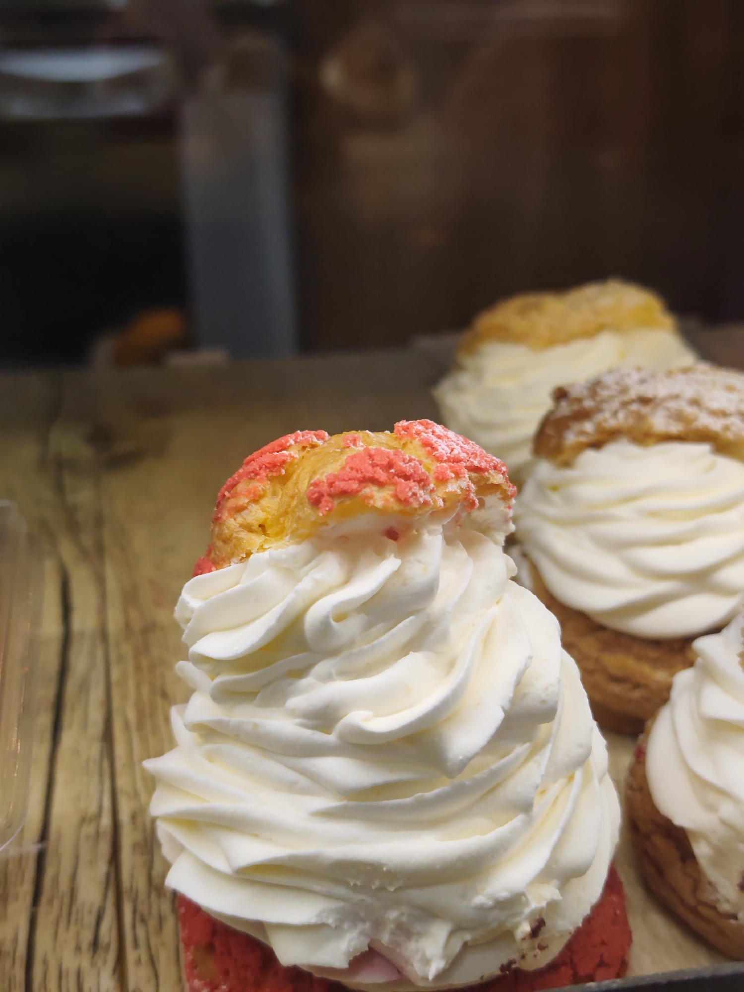 The image shows a close-up of several cream puffs on a wooden surface. The foreground puff is topped with a swirl of white cream and a piece of crunchy, pink-speckled pastry. In the background, other cream puffs are visible, some dusted with powdered sugar. The lighting suggests an indoor setting, possibly a bakery or display case. The scene does not contain any identifiable landmarks or elements that would suggest a location in Sochi, Russia.