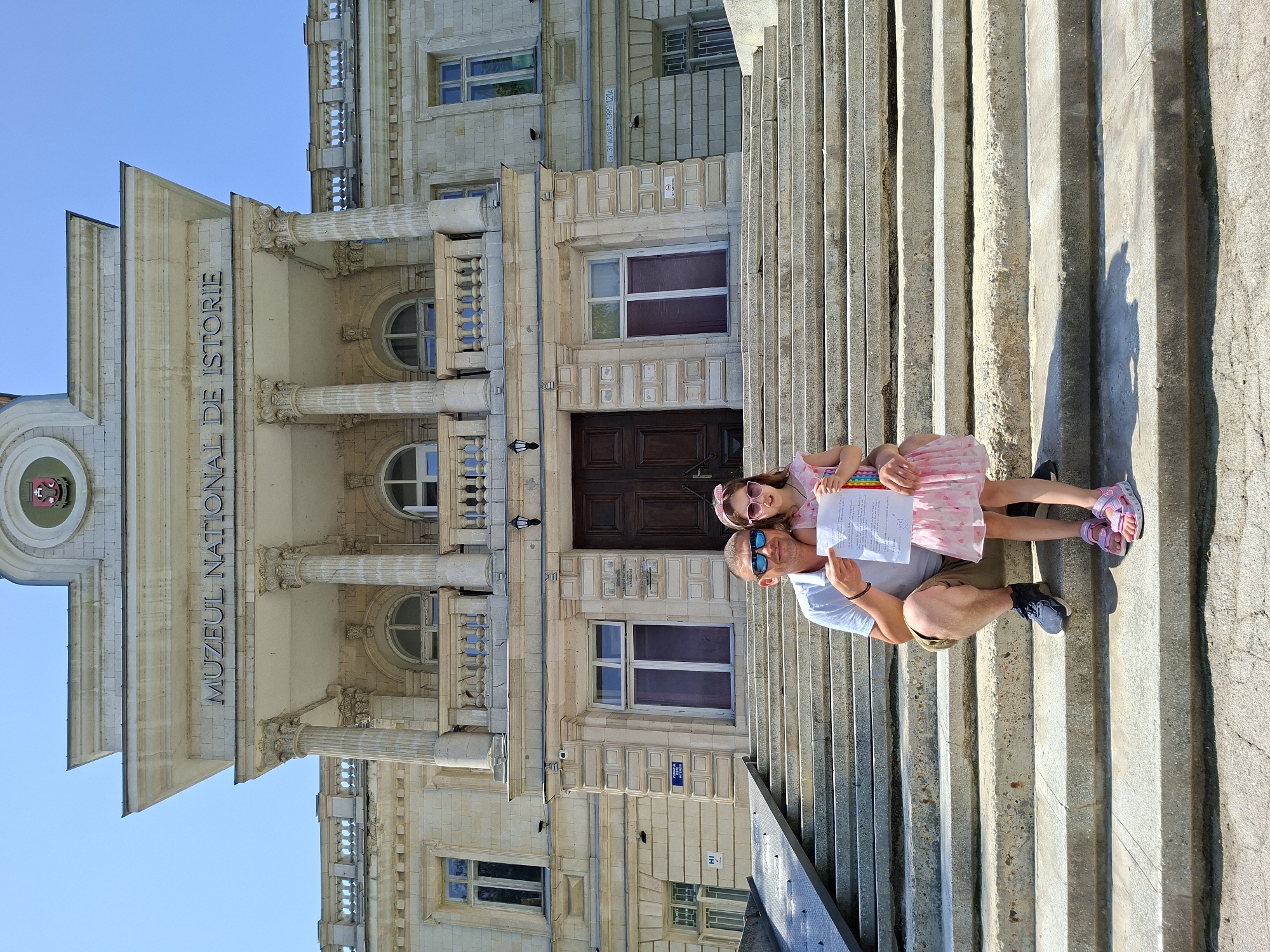 In front of the National Museum of History of Moldova in Chișinău, a man and a young girl are posing on a wide, stone staircase. The man, wearing a light blue t-shirt, khaki shorts, and dark sneakers, is kneeling on a step, holding a document and pointing at it with his right hand. He has sunglasses on and is looking at the camera. The girl, standing beside him, is wearing a pink dress, pink sunglasses, and pink sandals. She is holding a colorful, rainbow-striped object with her right hand. The museum building is made of stone, with large columns and ornate architectural details. The sky is clear and blue.
