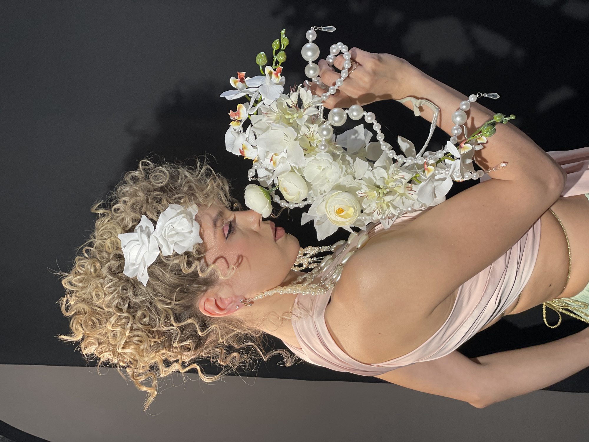A woman with blonde curly hair adorned with white flowers is positioned in front of a black backdrop. She is wearing a light pink dress and pearl jewelry. She holds a bouquet of white flowers and pearl strands. The image has a dark ambiance. It is unclear where the image was taken in Дачное, Russia.