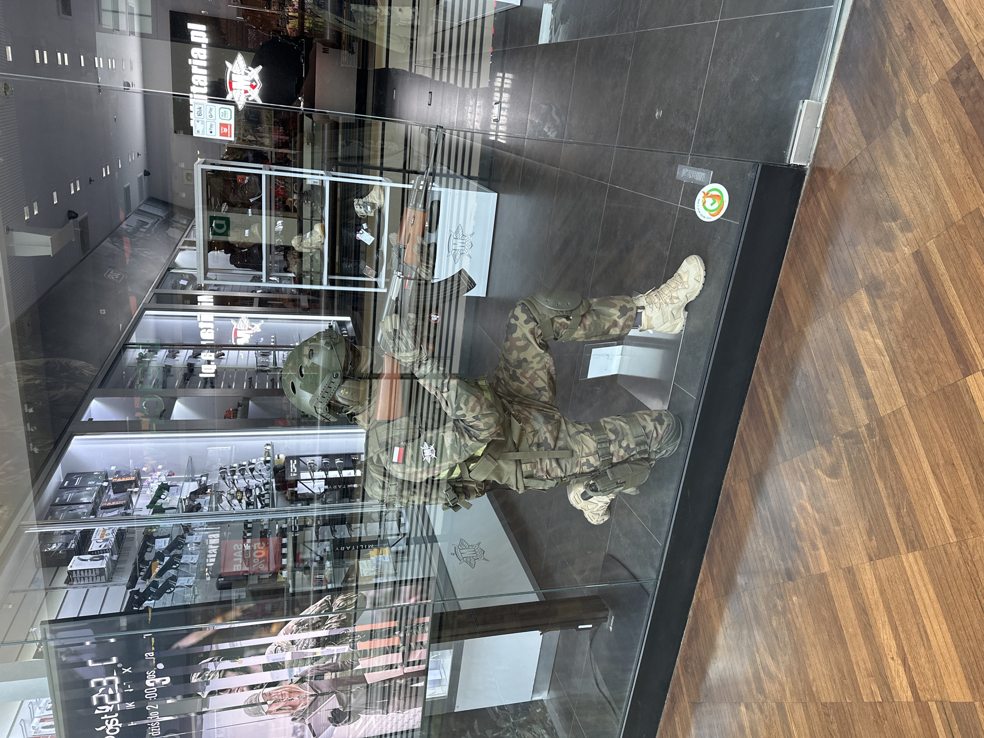 The image shows the storefront of a military surplus store, likely located in Łódź, Poland, given the context provided. The store's window display features a mannequin dressed in camouflage military attire, kneeling and holding a rifle. The mannequin is equipped with a helmet, tactical vest, and a holster on its leg. Behind the mannequin, shelves are visible displaying various military-style items, including what appear to be replica firearms, sunglasses, and other accessories. The store's signage, visible in the reflection on the glass, includes "militaria.pl," a website for a Polish military surplus retailer. The flooring inside the store appears to be dark tile, while the flooring outside is a polished wooden parquet. The scene is well-lit, suggesting it is daytime.