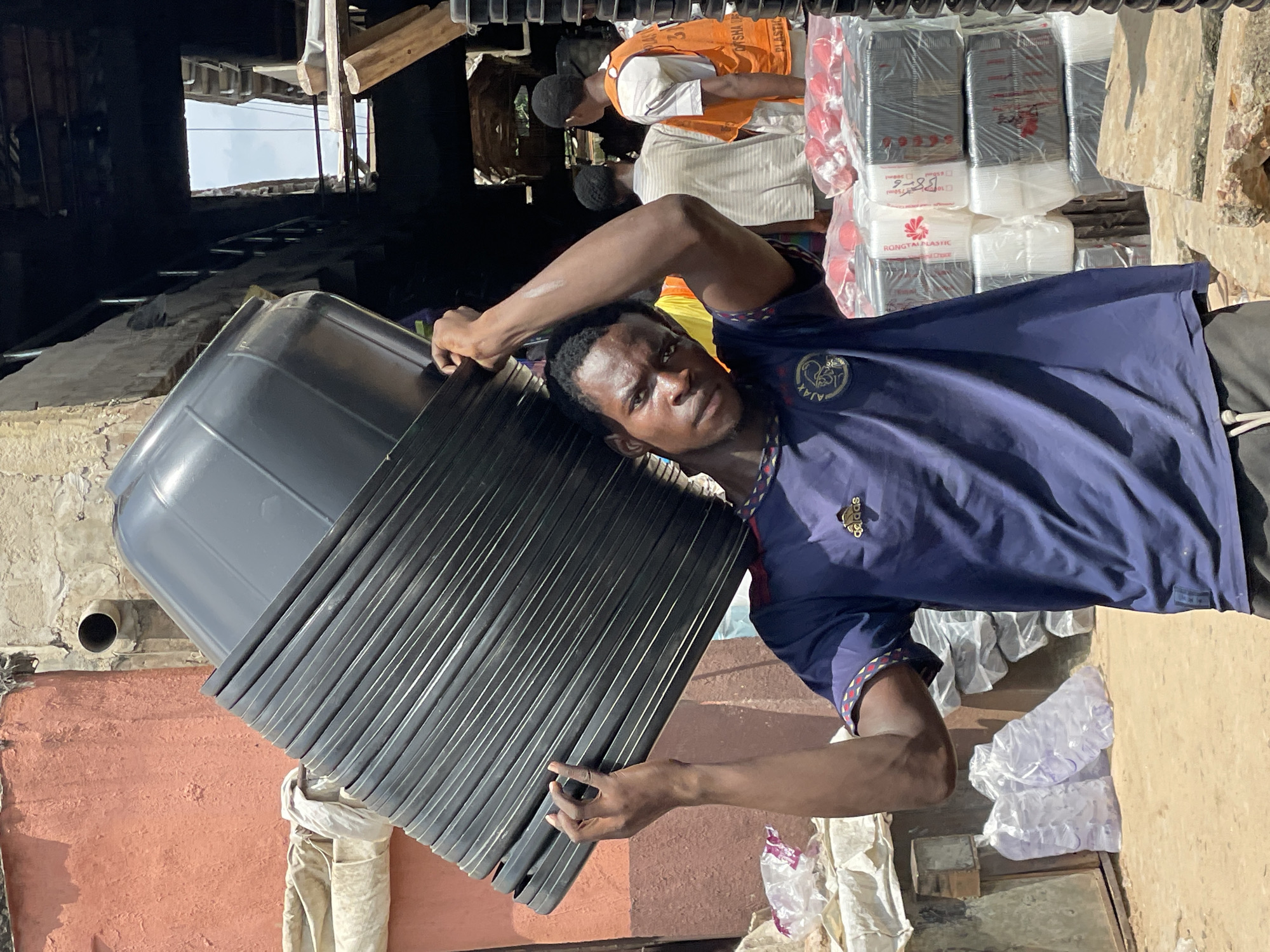 In an outdoor urban environment, a young Black man is carrying a large stack of black plastic tubs on his head. He is wearing a dark blue t-shirt with an Adidas logo and a crest on the left chest, and dark shorts. The tubs are stacked precariously high and appear to be a significant weight. The background shows a mix of buildings and market stalls. To the left, a textured pink wall with a grey pipe is visible. Behind the man, a market scene unfolds with people, stacks of goods wrapped in plastic, and wooden structures. Sunlight filters through gaps in the overhead structures, creating areas of light and shadow. The ground is covered in dirt and debris. Other individuals, including men wearing orange and white vests, are visible in the background, suggesting a commercial area.