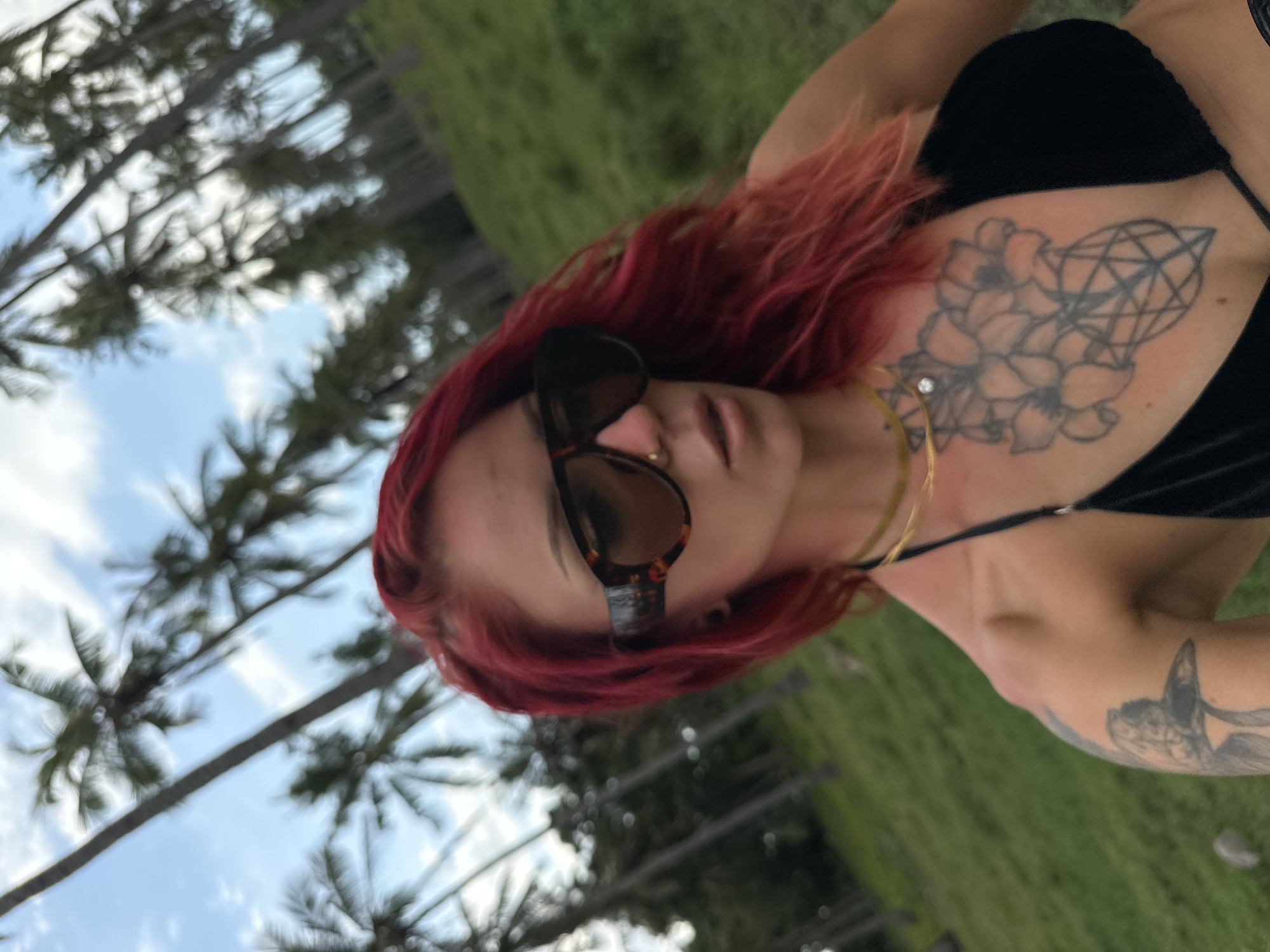 A woman with red hair, sunglasses, and a black bikini is pictured from a low angle. She has a tattoo on her chest and arm. Palm trees and sky are in the background, and green grass is visible. The location is Unknown, Indonesia.