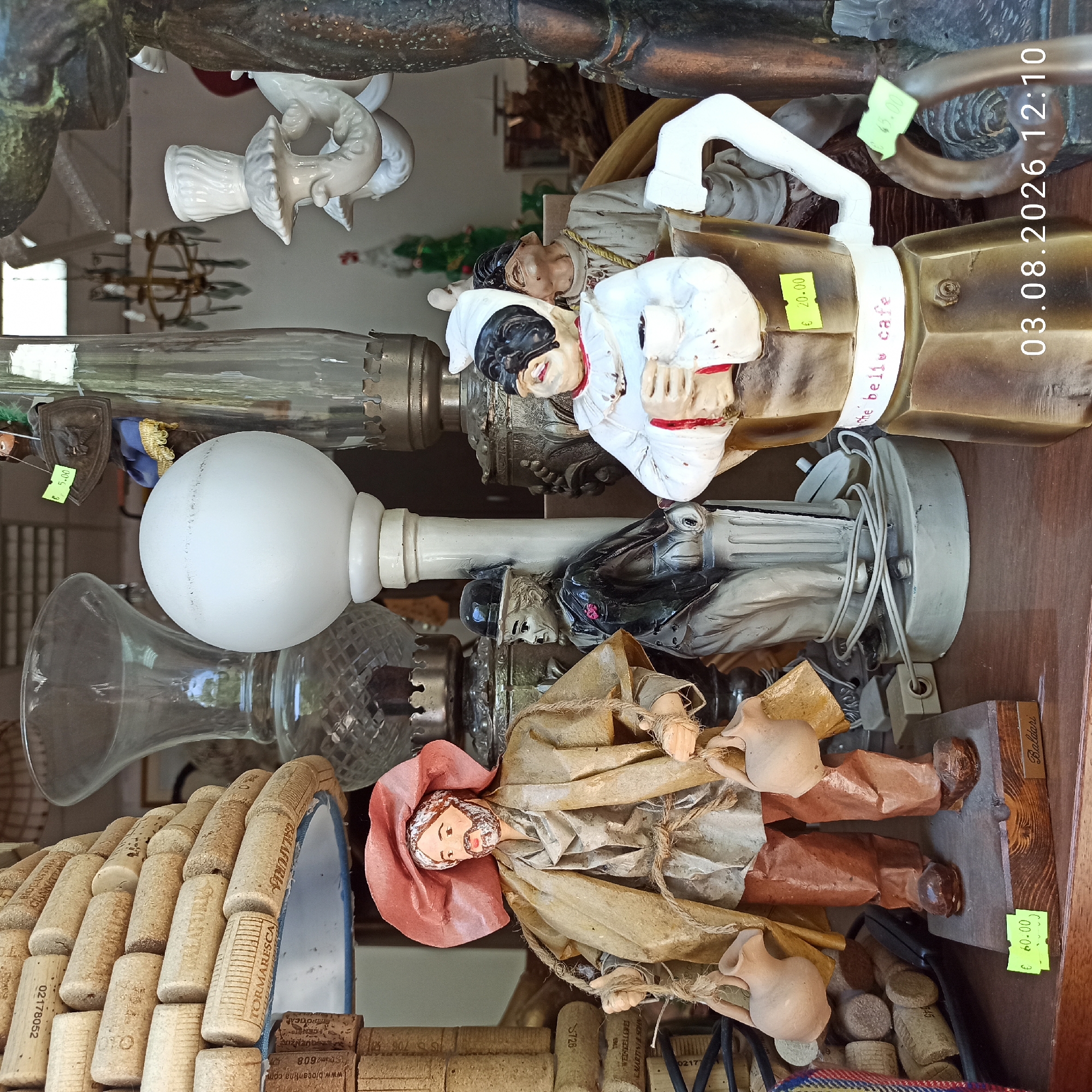 The image captures a close-up view of a collection of decorative items and figurines displayed on a wooden surface. The setting appears to be an indoor market or shop, possibly in Ruvo di Puglia, Italy, suggested by the presence of various artisan crafts.
In the foreground, a wooden figurine of a man with a straw hat and carrying two clay jugs is prominently featured. The figurine is made of textured material, possibly paper or fabric, and wears a beige cloak and brown pants. A price tag displaying "€ 60.00" is attached to its wooden base.
To the right of the man figurine, a ceramic figurine resembling a character from the Italian commedia dell'arte, possibly Arlecchino, is depicted. This figure wears a white costume with red trim and a black mask, holding a small white cup. A price tag of "€ 20.00" is affixed to its body. Behind this figure, another figurine with a similar mask and costume is partially visible.
Further to the right, a metal sculpture of a handle or part of a larger decorative piece is visible, with another price tag of "€ 15.00".
In the background, several glass lamps and other decorative objects are present. A large, clear glass vase with a diamond pattern is visible, as is a globe-shaped lamp with a white surface. Other elements include what appears to be a tower made of wine corks, stacked to form a conical shape. A portion of an ornate chandelier is also visible in the upper background.
The lighting suggests an indoor environment, possibly illuminated by natural light filtering in from an unseen source, and supplemented by artificial lighting. The overall impression is of a retail display of unique and handmade items, with a focus on traditional Italian crafts and decorative pieces. The date stamp "03.08.2026 12:10" in the lower right corner indicates the time the image was captured.