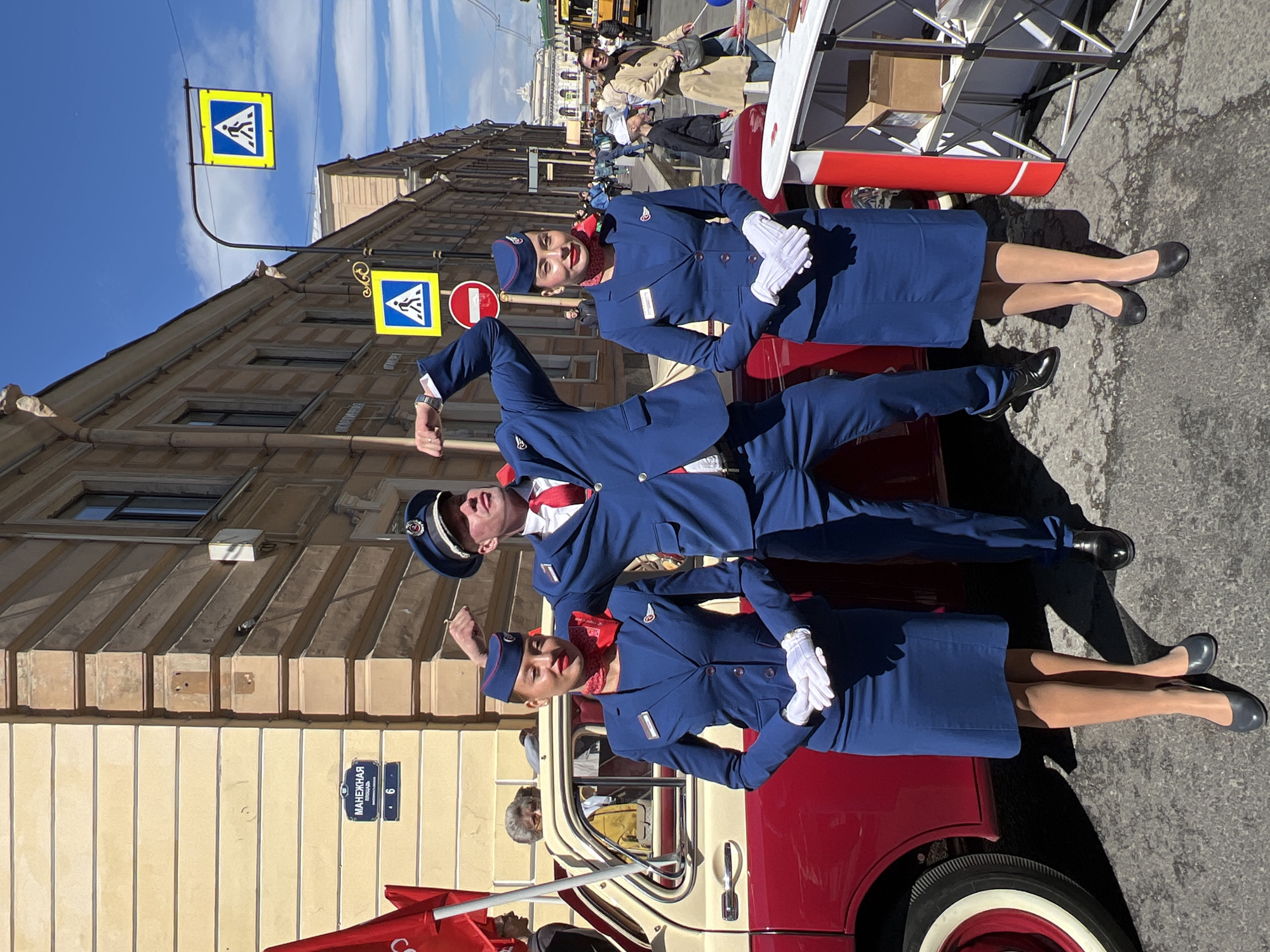 The image captures a scene in Saint Petersburg, Russia. Two women dressed in matching blue stewardess uniforms with white gloves and red neck scarves pose on either side of a man in a matching blue pilot's uniform, who stands on the hood of a vintage red and white car. The street sign indicates the location is "Manezhnaia street, building 6". Pedestrian crossing signs are visible on the building above. In the background, pedestrians and other vehicles populate the street, while a grand building is visible further down the street. The day is sunny with a blue sky.