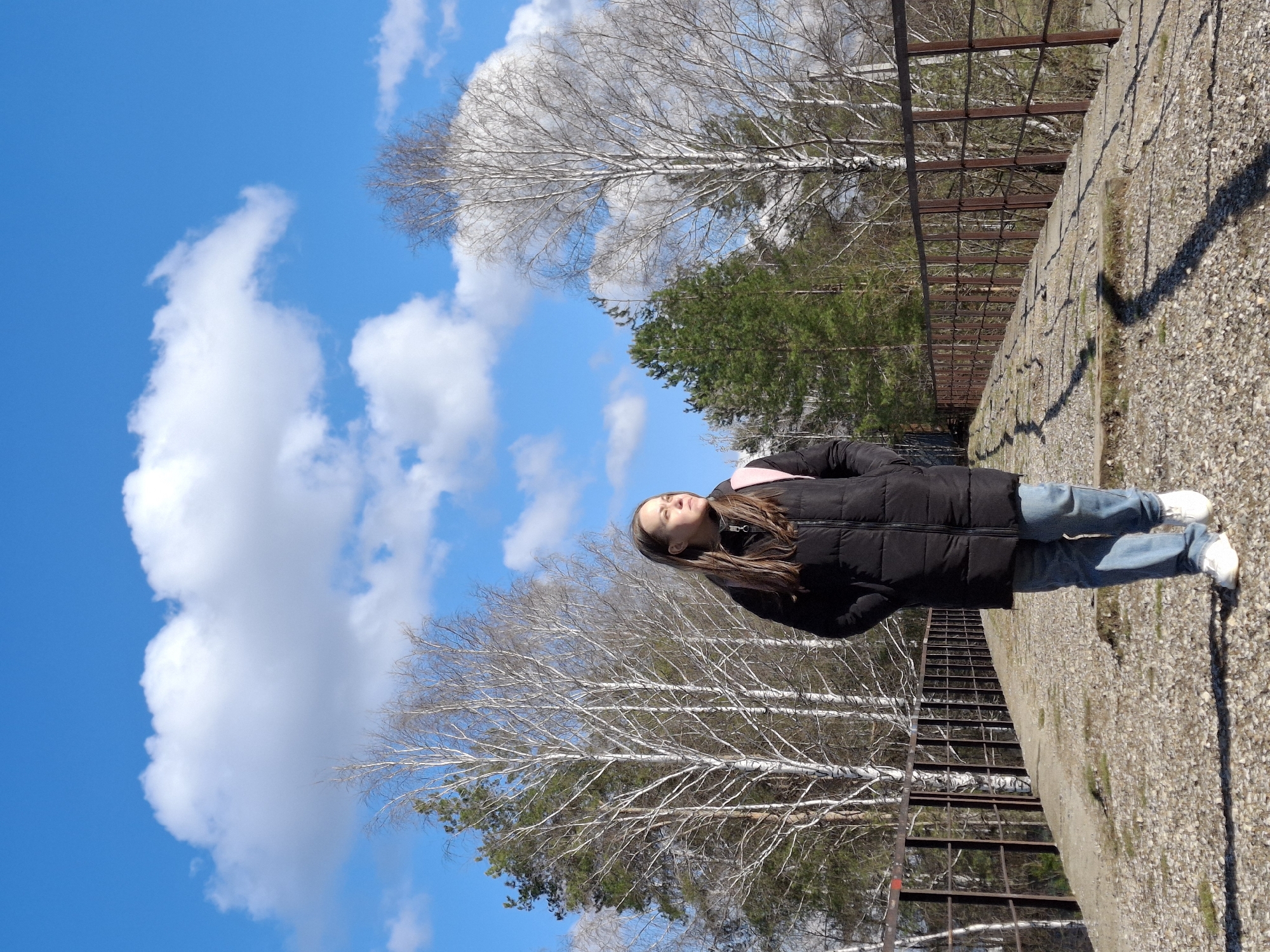 A young woman stands on a gravel surface, facing the sky. She wears a black puffer jacket, blue jeans, and white sneakers, with a pink backpack strap visible on her shoulder. Her long, brown hair flows down her back. She appears to be gazing upwards.
Behind her is a landscape of trees, including bare birch trees and evergreen pines, set against a bright blue sky with fluffy white clouds. A rusted metal fence runs along the edge of the gravel surface where she stands, possibly indicating a ledge or drop-off.
The gravel surface looks like a road. A long shadow stretches onto the road.
The location might be near the forest in the city of Chaikovsky, Russia.