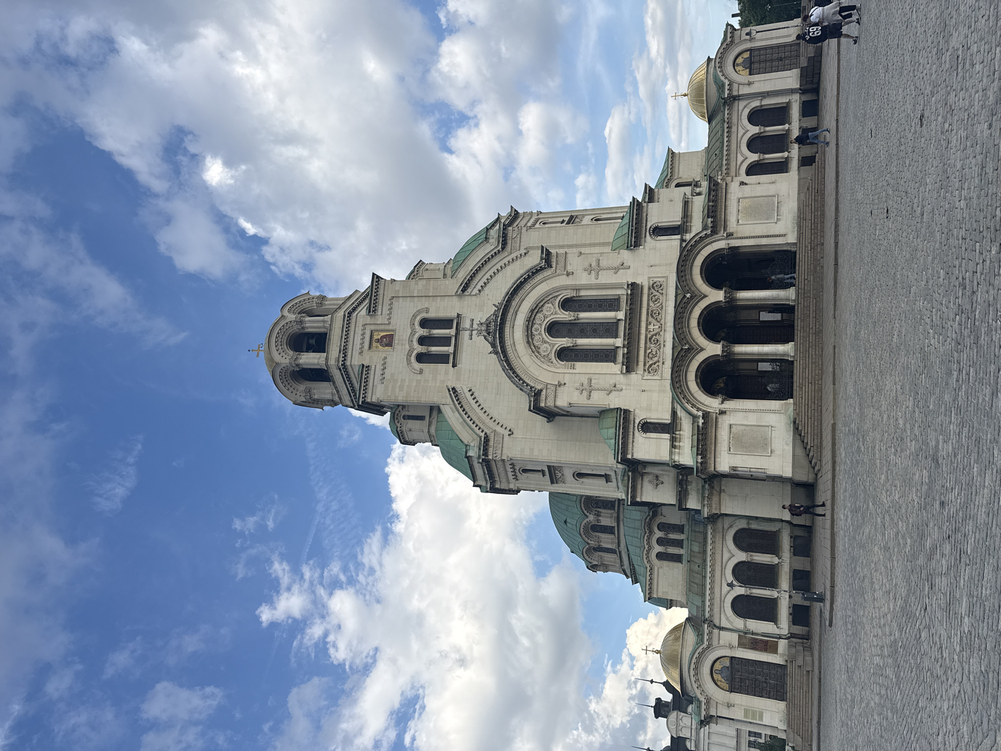 The image shows the Alexander Nevsky Cathedral in Sofia, Bulgaria, viewed from a low angle. The cathedral is a large, ornate building with white stone, green domes, and gold accents. The blue sky is visible behind the cathedral, filled with scattered clouds. A paved area is located in front of the cathedral. Two people are visible in the upper right corner of the image, and a person is walking near the cathedral entrance.