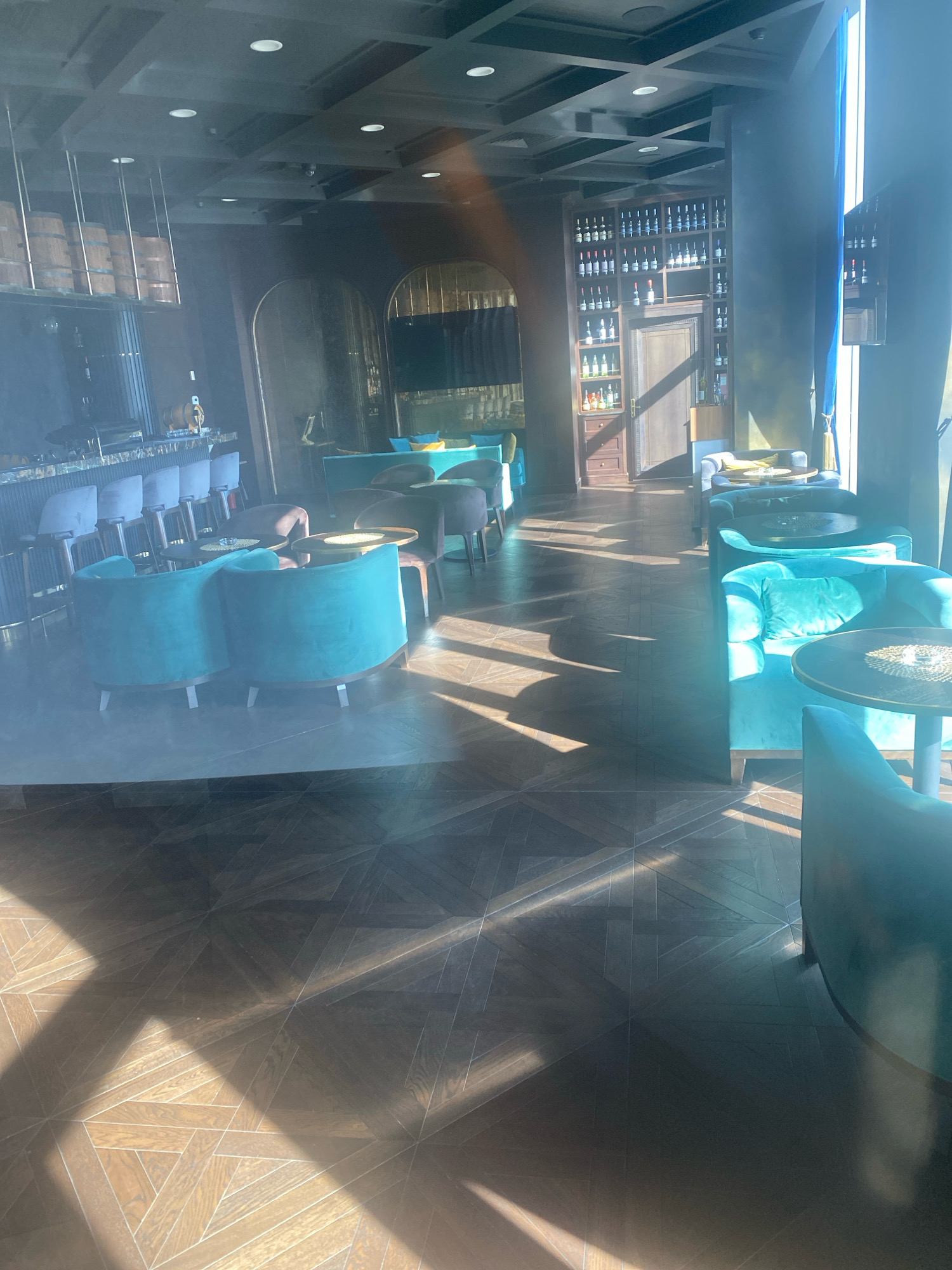 The image depicts the interior of a dimly lit, upscale establishment, likely a bar or restaurant, with strong natural light streaming in from the right, casting angular shadows across the dark wooden floor. In the foreground, two plush, teal-colored armchairs are positioned side-by-side, facing away from the viewer. To the right, a small, round table with a metallic top sits next to another teal armchair. Further back, a row of similar teal armchairs and chairs are arranged around small, dark tables.
Along the left side of the frame, a bar area is visible with a polished counter and several bar stools. Above the bar, a rack holds a collection of wooden barrels. A wall unit filled with bottles of wine dominates the background on the right, with a dark wooden door in the center of the unit. The ceiling features an intricate dark grid pattern with recessed lighting. The overall ambiance suggests a sophisticated and contemporary setting.
Based on the visual cues, this establishment is identified as "The Loft" bar located in the city of Minsk, Belarus.