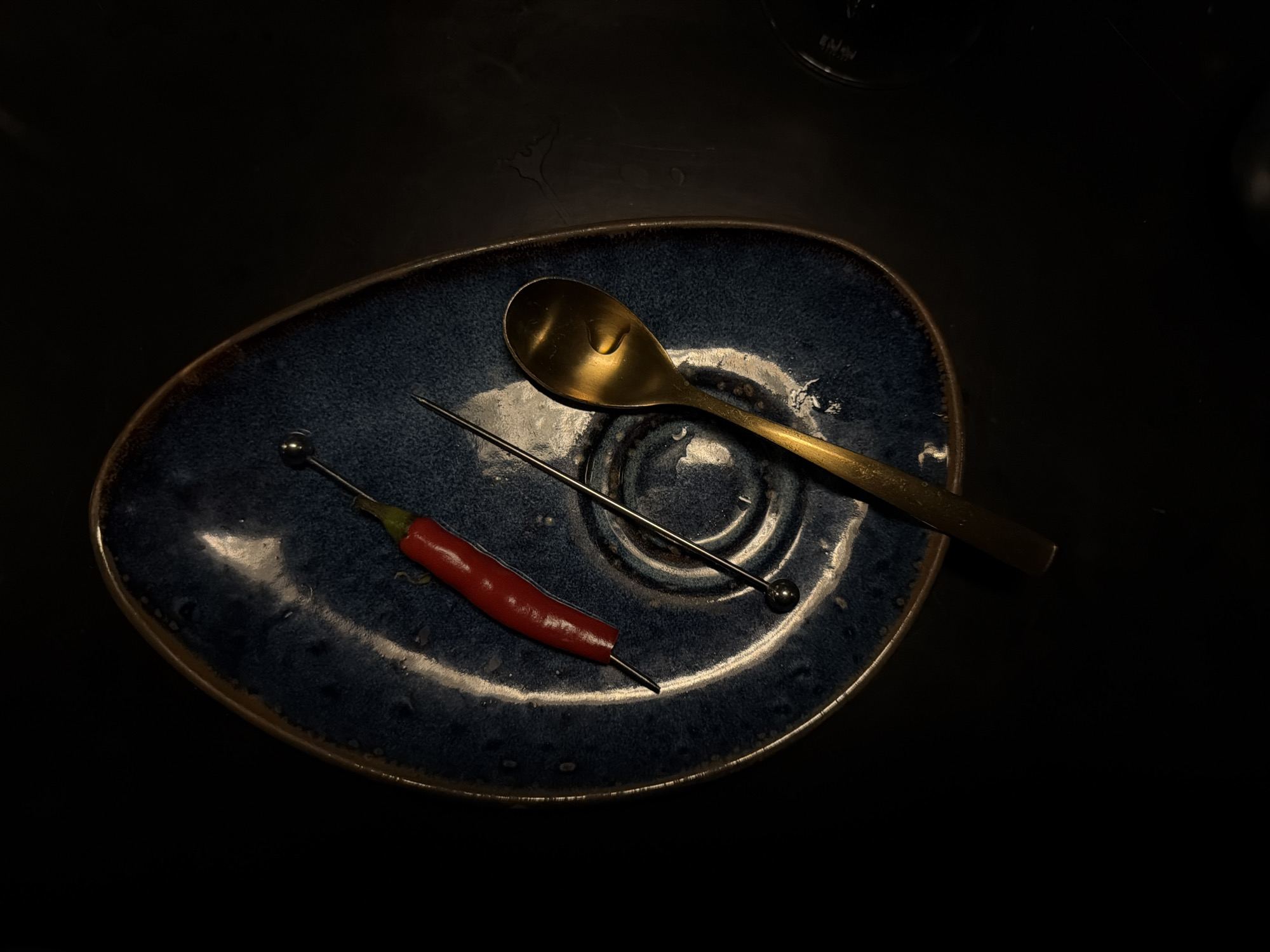 A top-down, darkly lit photograph showcases a minimalist composition centered on an irregularly shaped, deep blue ceramic plate. The plate, with its textured, mottled glaze and brown rim, rests on an undefined dark surface, possibly a table, which displays subtle reflections and faint outlines of moisture or wear.
Arranged on the plate are three primary objects: a golden-colored metal spoon, a slender silver-colored metal cocktail pick, and a vibrant red chili pepper. The spoon, positioned towards the top right of the plate, gleams under the light and appears to hold a small droplet of liquid in its bowl. Below it, the cocktail pick extends across the plate, featuring a small spherical detail at one end and a sharp point at the other, which appears to be inserted into the stem-end of the chili. The whole red chili pepper lies horizontally on the bottom left side of the plate, its smooth, curved form contrasting with the rugged texture of the ceramic.
The lighting is dramatic and focused, highlighting the objects on the plate while keeping the surroundings in deep shadow, creating a moody and sophisticated atmosphere. In the blurry top right background, a partial view of a glass or bottle is visible, upon which barely legible text, possibly "ENJOY," can be discerned. No direct activity is depicted; the image presents a carefully composed still life, potentially suggesting elements for a drink or appetizer. The scene appears to be indoors, likely in a restaurant or bar setting, under low light conditions.