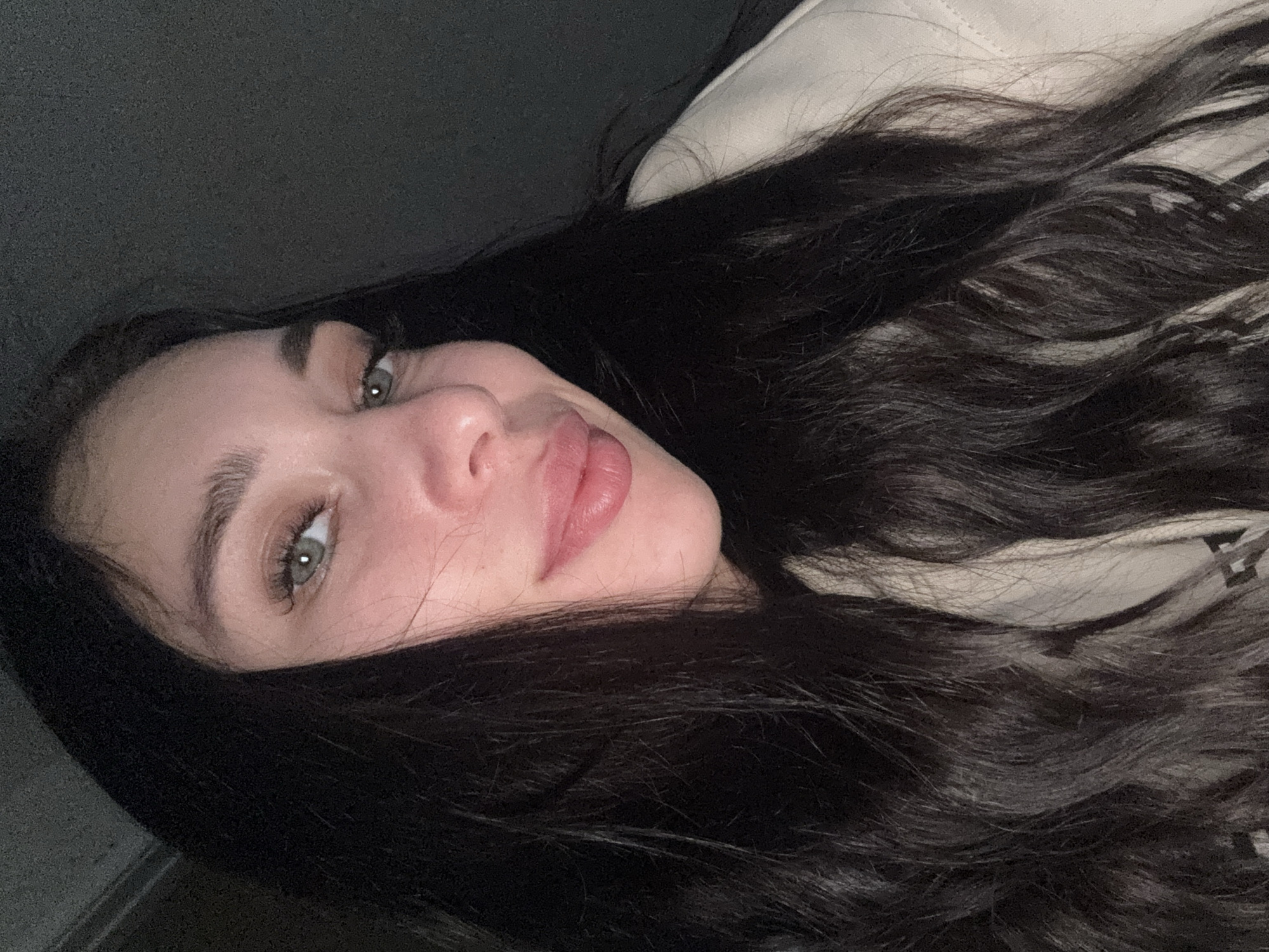 The media file presents a close-up portrait of a young woman, captured in a sideways orientation, suggesting she is lying down or the camera was held horizontally. She gazes directly towards the viewer with a gentle, soft smile. Her head is tilted, showcasing her facial features prominently.
She has long, dark, wavy hair that frames her face and spreads across the frame, partially resting on a light-colored, possibly beige or off-white, garment that appears to be made of a soft fabric, like a hoodie or jacket. Her eyes are a striking green or hazel, with well-defined eyebrows and visible eyelashes. Her lips are full and have a subtle pink hue, showing a slight sheen. Her skin appears clear and well-lit.
The background is dark and largely indistinct, suggesting an indoor setting with subdued, soft lighting that primarily illuminates her face. A darker, textured surface is visible on the left side of the frame, possibly a wall or furniture. There is no visible text in the image. This photograph is set in Chaikovsky, Russia.