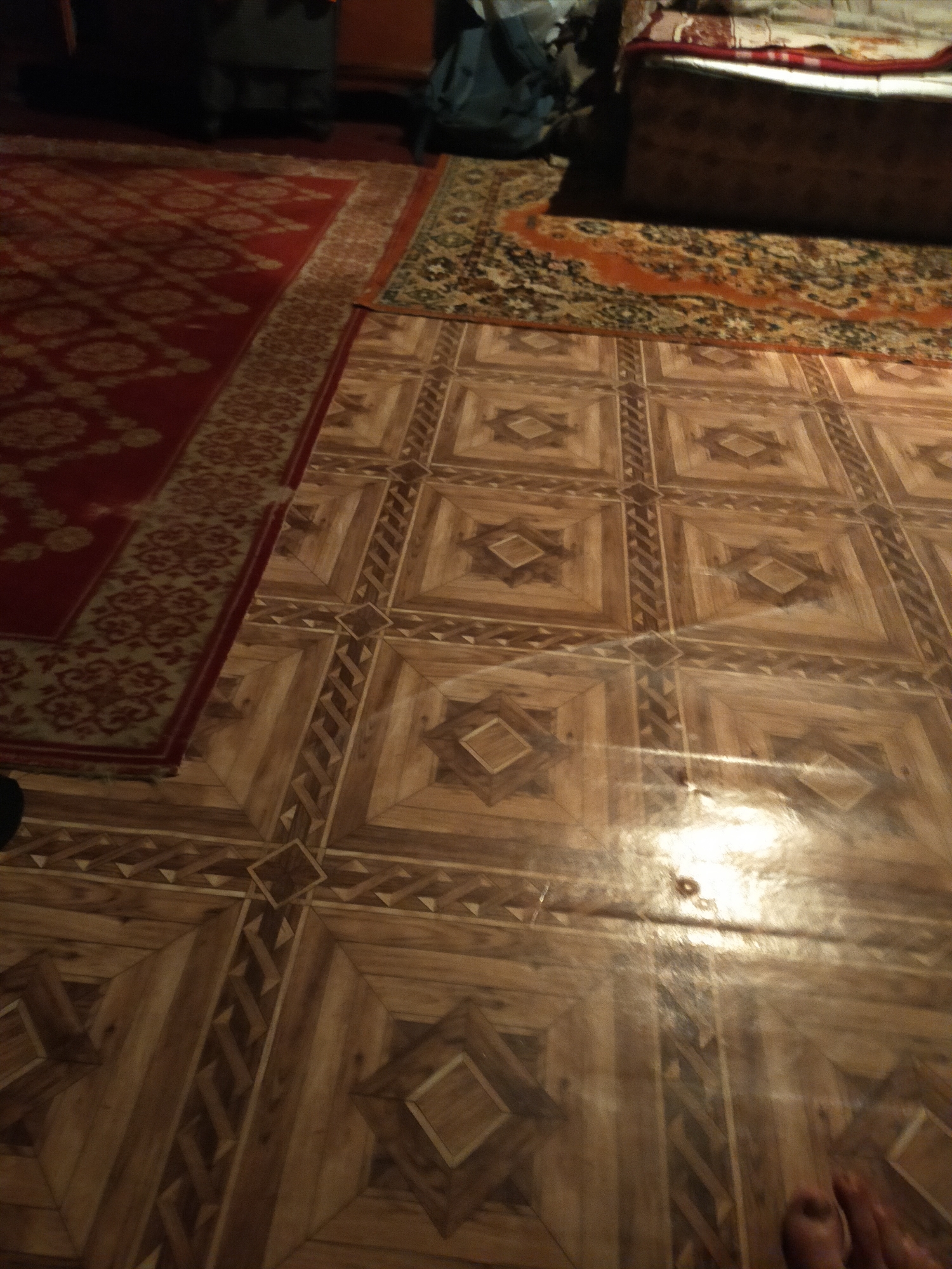 The image captures an indoor scene within a room in the city of Unknown, Unknown. The floor is covered by multiple distinct materials. The predominant surface in the foreground and center is a glossy, patterned linoleum or laminate flooring, designed to resemble parquet wood. This flooring features a grid of squares, each containing a central diamond motif, and connecting strips with geometric patterns in various shades of brown and tan. A prominent, bright, oblong reflection from an unseen light source is visible on this surface.
To the left, a large red rug with cream and gold floral and medallion patterns is partially visible. This red rug overlaps a narrower, cream and brown patterned rug that serves as a transition to the wood-look floor. In the upper right quadrant, a section of furniture, possibly a bed or divan, is visible, covered with red, white, and brown patterned textiles. This furniture rests on another patterned rug, primarily green and cream. The upper left portion of the frame is largely obscured by shadow, showing indistinct dark furniture legs and a dark blue bag. A portion of a human foot, specifically the toes, is visible at the extreme bottom right corner of the frame.