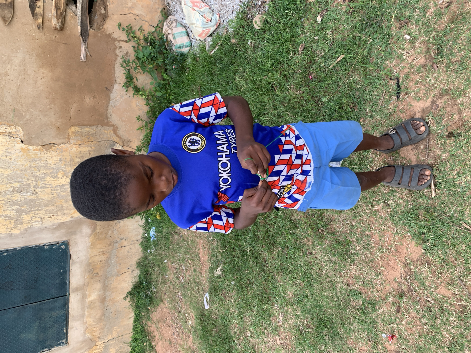 A young Black boy stands on a patch of grass and dirt outside a dilapidated building. He is wearing a blue Chelsea FC jersey with the Yokohama Tyres logo and blue shorts. His hands are busy manipulating a green string. His feet are clad in dark grey sandals. The building behind him is made of weathered tan plaster with cracks and peeling paint. A dark green, diamond-patterned metal sheet is affixed to the wall to the left, resembling a small window or vent. To the right, the wall shows a hole with some wooden debris protruding. Several bags, possibly cement or fertilizer, are piled on the ground near the building. The ground is a mix of sparse green grass, dry brown patches, and loose dirt. Scattered pieces of white and colored debris are visible on the ground.