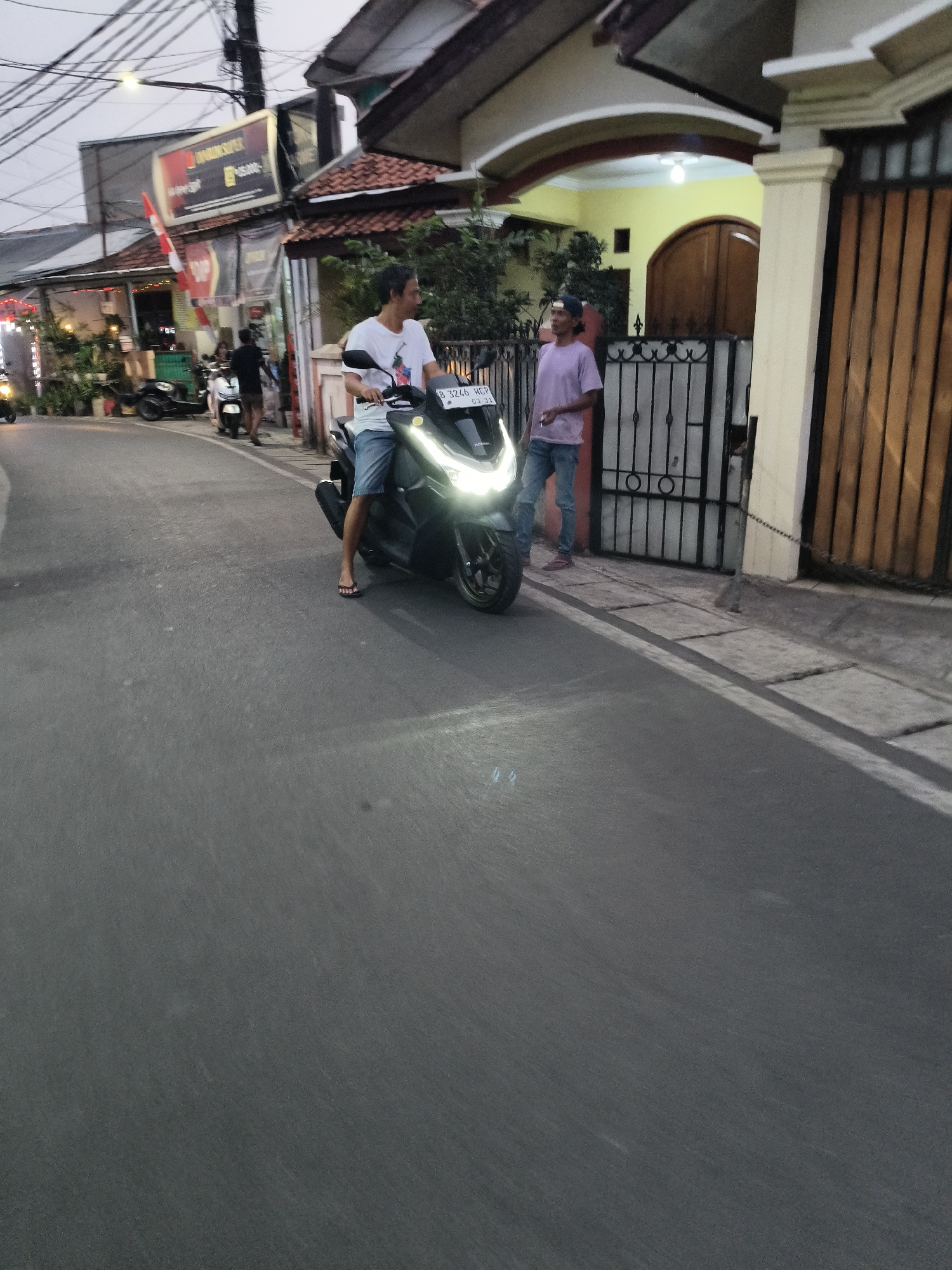 A young man is seated on a black motorcycle on a street in South Tangerang, Indonesia. He wears a white t-shirt and denim shorts, and his feet are bare, clad in flip-flops. His right hand rests on the handlebars. Beside him, another young man stands on the sidewalk. He is dressed in a light purple t-shirt and blue jeans, with a baseball cap on his head. He holds a cigarette in his left hand. The motorcycle's headlights are on, illuminating the road. In the background, a house with a yellow wall and wooden doors is visible, as well as a metal gate. The street is paved, and there are buildings lining the side of the road, some with signs. The sky is overcast, suggesting it may be late afternoon or evening.