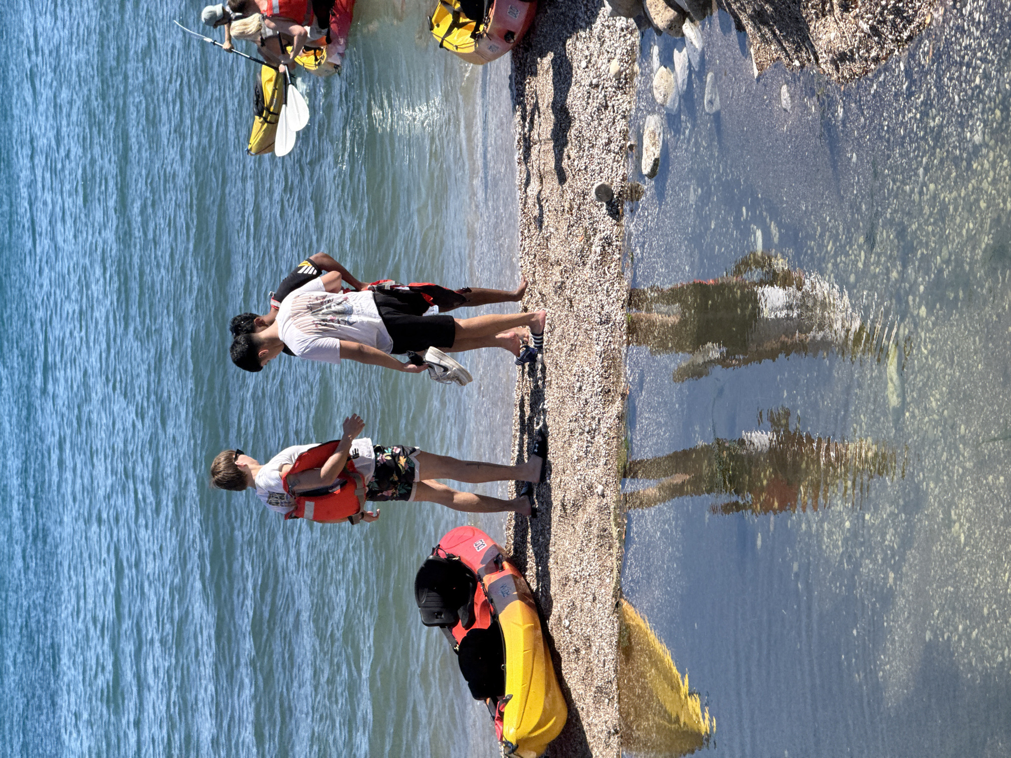 The image depicts a beach scene near the city of Budva, Montenegro. In the foreground, a calm body of water reflects the sky and the figures of people standing on a pebble beach. Two kayaks are visible on the shore, one yellow and red, the other red. In the background, three young men are standing near the water's edge. One is wearing a red life vest and floral patterned shorts, another a white t-shirt and black shorts, and the third a black and white striped shirt and red shorts. Two of the men are looking at each other, while the third is looking down. Further out in the water, another kayak with two people is visible. The water is blue and appears to be the Adriatic Sea. The beach is composed of small pebbles and rocks.