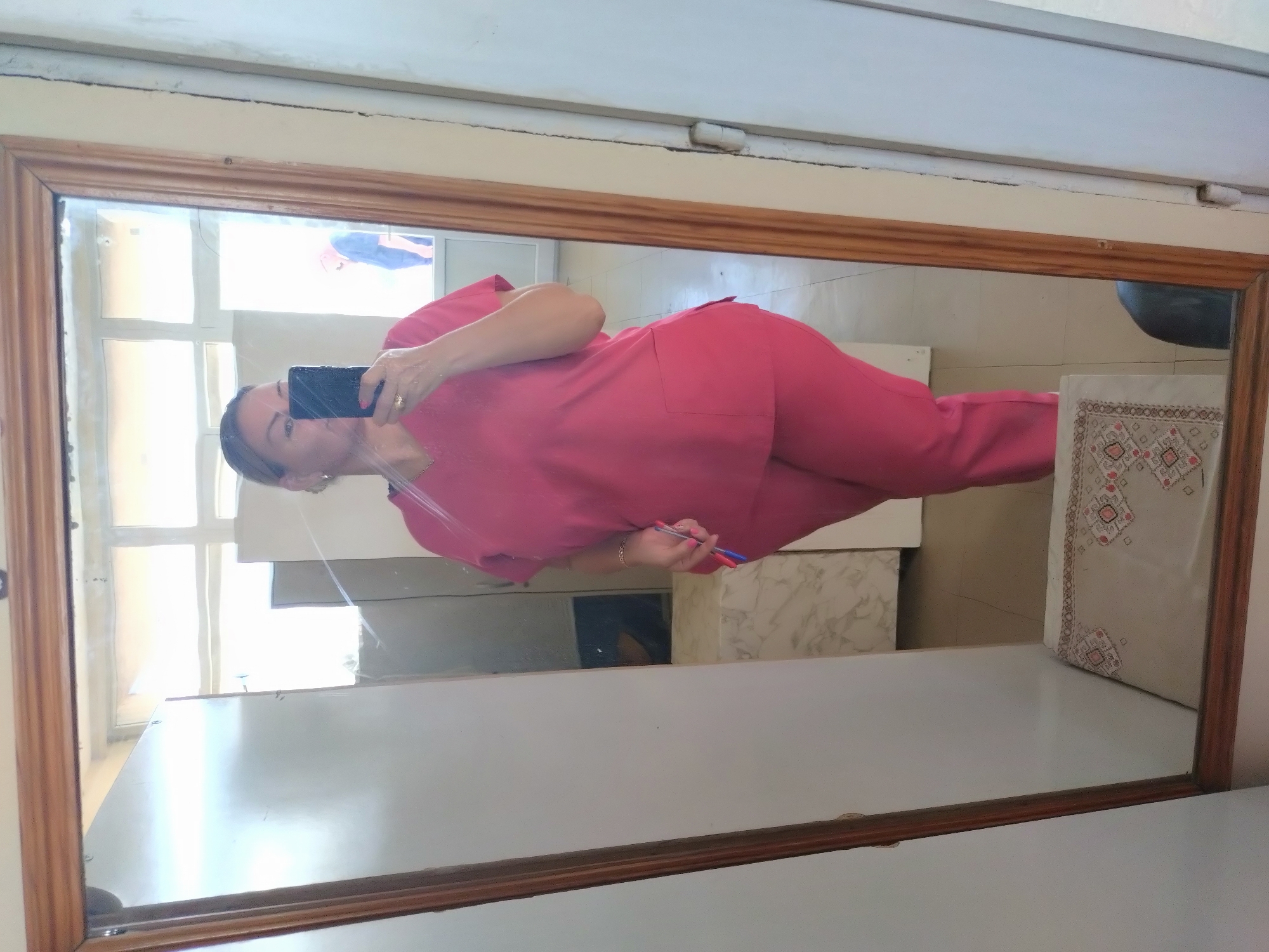 A woman in pink scrubs is taking a selfie in a mirror. The mirror has a wooden frame and reflects a portion of what appears to be a room, including a window, a white surface, a patterned pillow, and a black object. The woman is holding a smartphone in one hand and a pen in the other. The location appears to be indoors, potentially in a medical facility in Stara Zagora, Bulgaria, given the scrub attire.