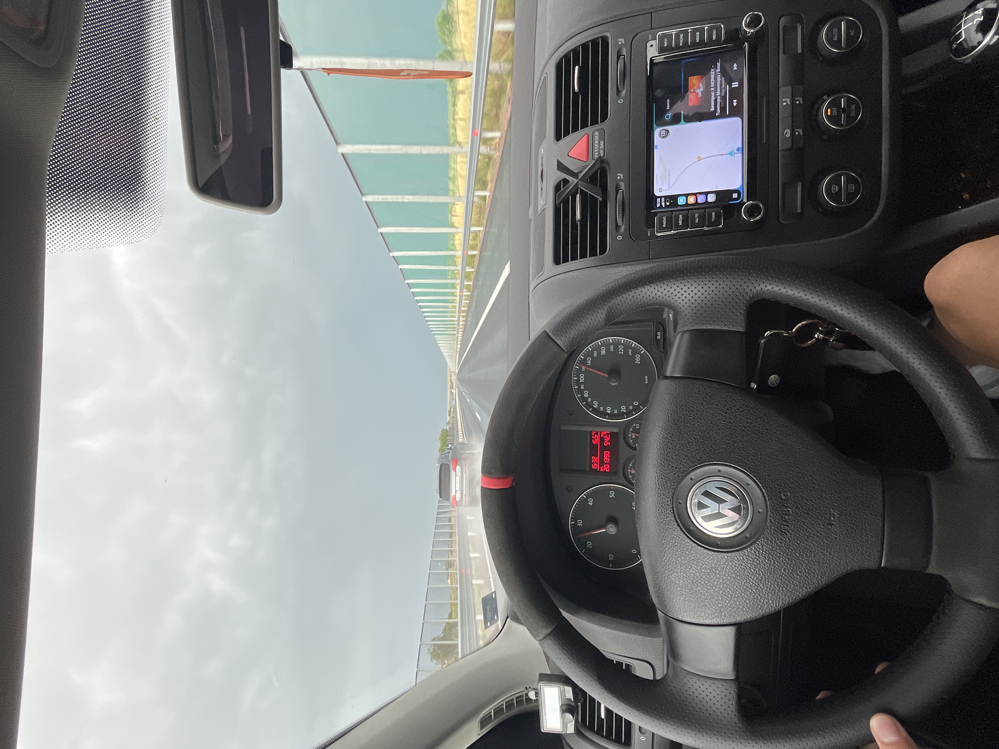 An internal view from a Volkswagen car shows the driver's perspective on a highway near Chokoba, Bulgaria. The image, taken during the mid-afternoon with an overcast sky, captures the dashboard, steering wheel, infotainment system, and the road ahead through the windshield.
In the foreground, a black, perforated leather steering wheel with a red accent stripe is visible, featuring the Volkswagen logo and "AIRBAG" text. The driver's right hand or arm is partially visible on the bottom right, suggesting the car is being driven. Behind the steering wheel, the instrument cluster shows a speedometer indicating a speed between 120 and 130 km/h, a tachometer, and a digital display with "15:32" (time), "16.5" (likely temperature in Celsius), "20", "1390", and "5427" (possibly trip or total mileage).
The center console features an infotainment system displaying a navigation map alongside music playback details, "Беатрис X MONKEY - Тинтири Минтири / Beat....". Buttons around the screen include "MENU", "PHONE", "NAV", "RADIO", "MEDIA", "MUTE", and "TONE". Below these are climate controls with "OFF", "DUAL", "AUTO", and "AC" buttons, and an "PASSENGER AIR BAG" indicator. A set of car keys hangs from the ignition or a nearby hook.
Outside the car, the highway extends into the distance under a grey, cloudy sky. The road appears damp or recently wet. A white sedan is visible some distance ahead. To the left, a sound barrier or guardrail with translucent green panels runs along the side of the highway, consistent with infrastructure found on European motorways. A rearview mirror is also visible at the top of the windshield. The overall scene depicts a neutral travel moment on a highway.