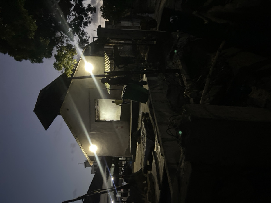 The image, captured at dusk or night, depicts an outdoor scene in Bagamoyo, Tanzania, dominated by a simple, light-colored building under artificial illumination. The sky is a deep twilight blue with visible clouds, suggesting early evening.
A prominent single-story building with a dark, corrugated metal roof stands on the left. Two bright outdoor light fixtures are attached to its facade, casting strong, ray-like beams across the scene and illuminating the building's exterior. Inside one of its window openings, which is brightly lit from within, a person wearing a light-colored (possibly yellow) shirt is standing, looking outwards or performing an activity.
To the right of the building, near one of the light sources, another figure is partially visible standing beside a green trash bin. The immediate surroundings of the building appear somewhat informal, with concrete walls, steps, and what seems to be debris or informal structures in the foreground and dark areas. Silhouettes of trees are visible against the sky in the upper portion of the image. The ground is uneven, characterized by various surfaces and possible rubble. No specific event beyond the presence of people in their daily environment is clearly discernable, and the atmosphere appears neutral. There is no visible text in the image.