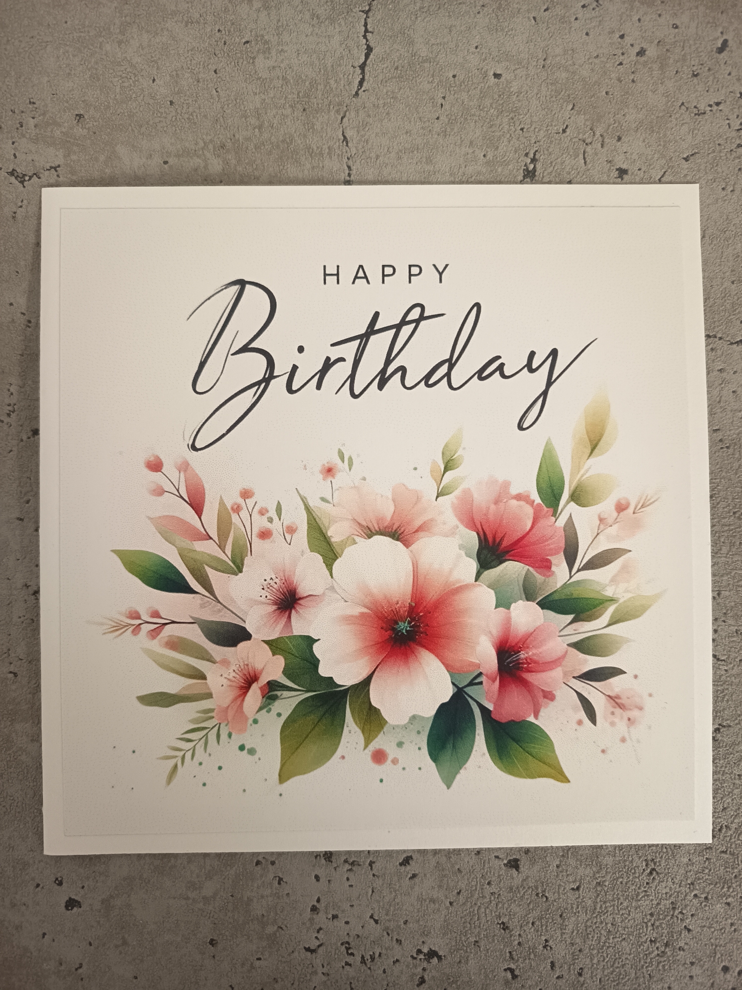 The image shows a "Happy Birthday" greeting card resting on a gray, textured surface. The card is square and has a white background with a border. The words "Happy Birthday" are printed in black font at the top. Below the text is a floral arrangement of pink and white flowers with green leaves. The floral arrangement appears to be watercolor-style art. There is no visible location in Wangen bei Olten, Switzerland.