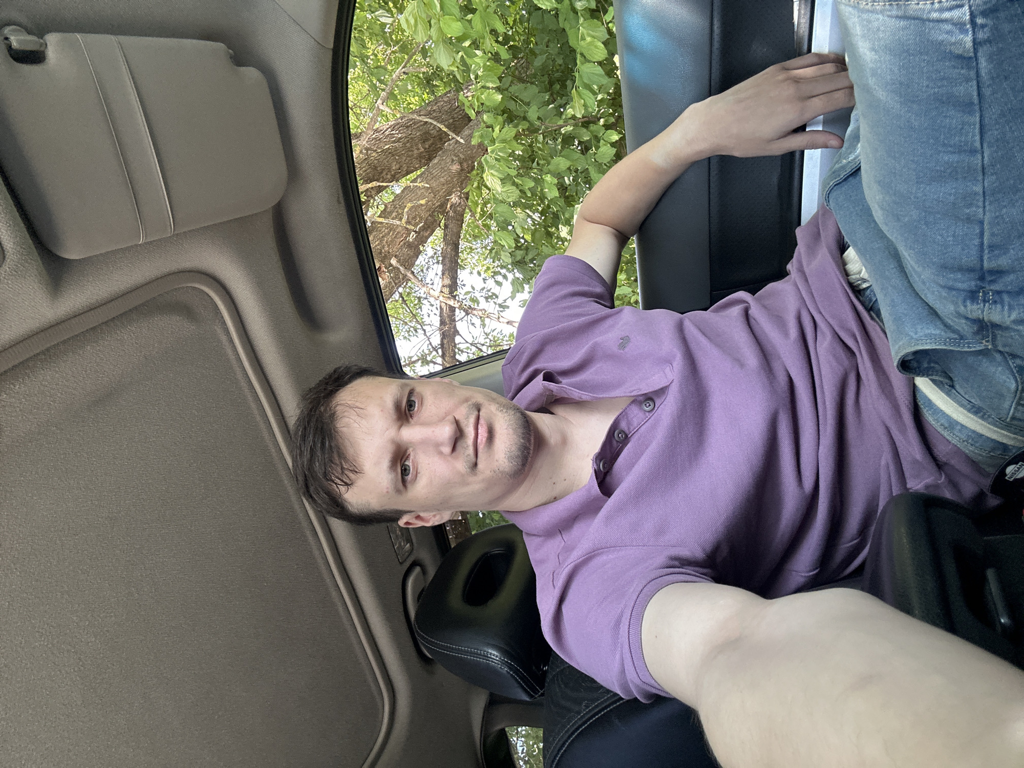 A man is sitting in the driver's seat of a car, taking a selfie. He is wearing a purple polo shirt and blue jeans. The car's sunroof is open, and trees and greenery are visible through the open window. The interior of the car, including the dashboard and seats, is visible. The man has short brown hair and stubble on his chin. He is looking directly at the camera with a neutral expression. The image was taken in the city of Makiv, Ukraine.
