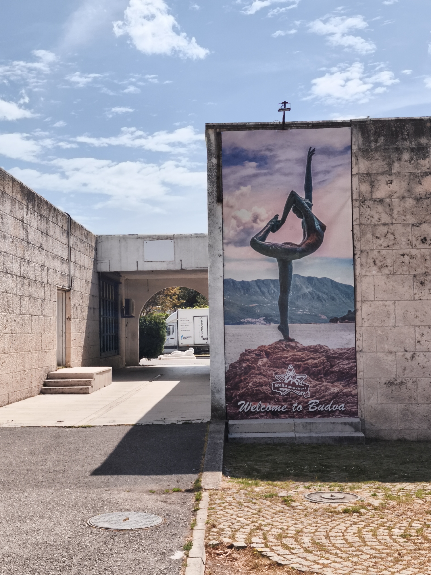 The image captures a street scene in Budva, Montenegro. On the right side of the frame, a tall, rectangular poster is affixed to a stone building, showcasing an image of a bronze sculpture of a ballet dancer on a rock outcrop near the sea. The poster bears the text "Welcome to Budva" and a logo, likely belonging to a local business. A cross-shaped antenna sits atop the building. To the left, a stone tunnel or archway leads further into the scene, with a vehicle visible in the background. The ground is paved with asphalt, transitioning to patterned stone blocks near the building on the right. The sky above is partially cloudy.