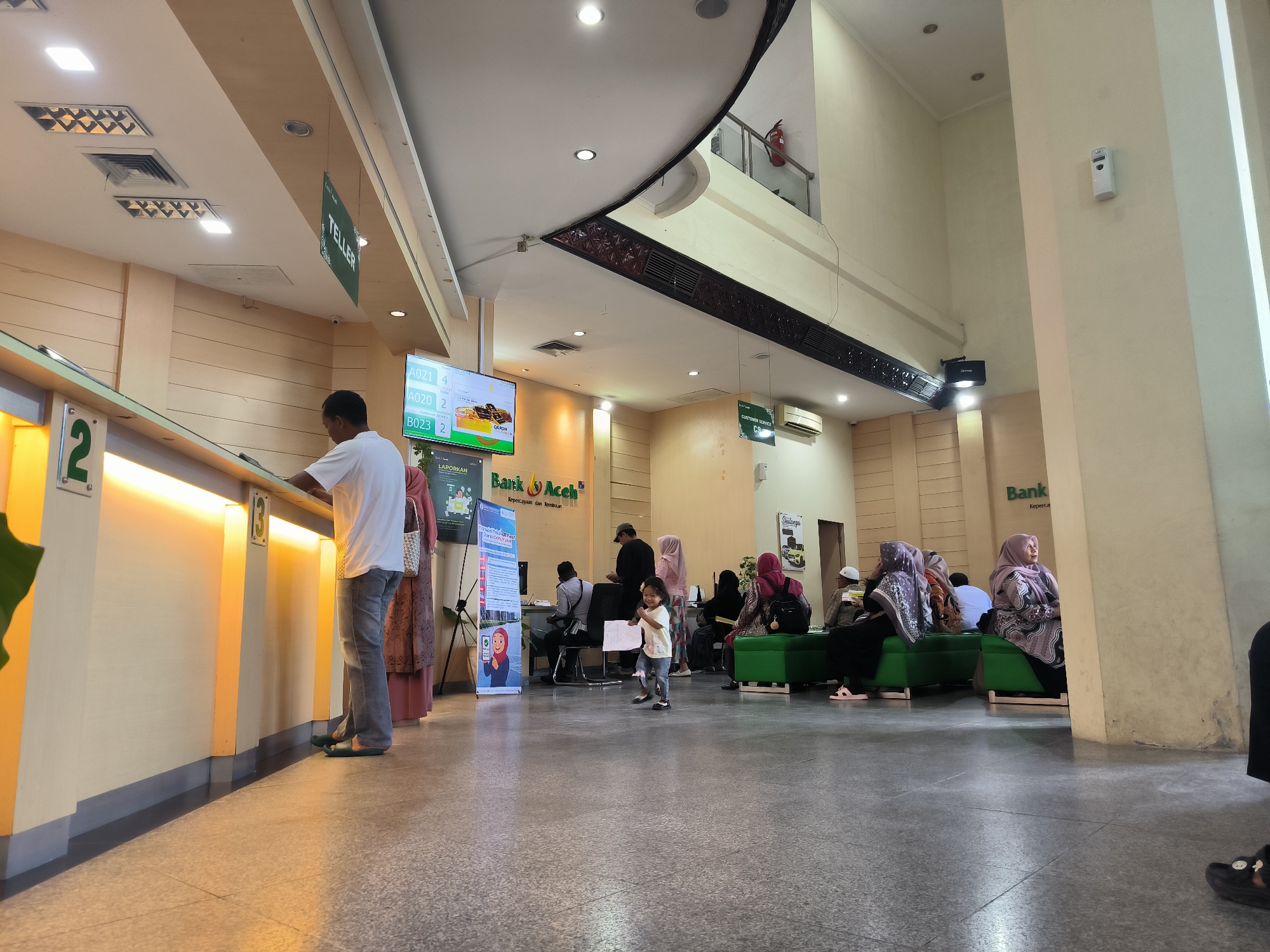 The image shows the interior of a Bank Aceh branch in Lhokseumawe, Indonesia. The scene is a bank waiting area. The floor is gray tile. A man in a white shirt and gray pants is standing at a teller counter marked with a "3". There is a green "TELLER" sign hanging above the counter. A woman in a brown dress is standing nearby. A young girl is walking in the middle of the waiting area, holding a paper. Several people, primarily women wearing hijabs, are seated on green benches. A "CUSTOMER SERVICE" sign is visible on the wall near a seating area. A display screen shows queuing numbers (A021, A020, B023). There is also a "Bank Aceh" sign, and promotional posters on stands. A man in a black cap is sitting down.