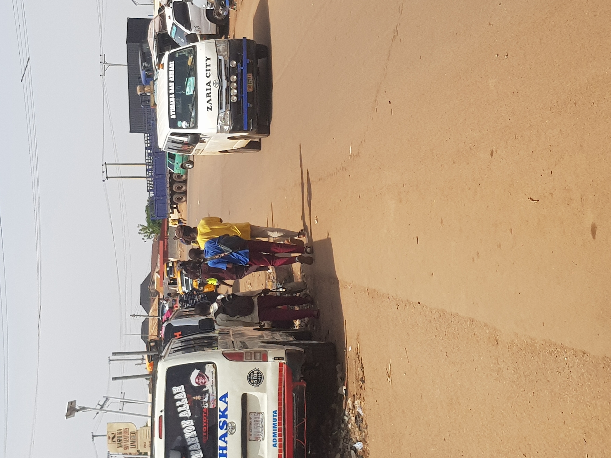 The image captures a bustling street scene in Kufena, Nigeria, on a clear, sunny day. The perspective is from the roadside, looking down a street filled with vehicles and pedestrians. The road surface appears to be a mix of asphalt and reddish-brown dirt.
In the foreground, on the right side of the road, several people are walking. One person in a yellow top and dark pants walks away from the viewer, while another in a blue shirt and dark pants follows. Shadows stretching from the people and vehicles suggest it is daytime, possibly morning or afternoon.
The road is lined with various vehicles. A prominent white commercial van, possibly a Toyota Hiace, is seen from the rear on the left side of the frame. Its rear window features text including "MANZON ALLAH" and a "TOYOTA" logo, along with "HASKA" in blue letters on its tailgate. A license plate "598XS" is visible, and the bumper has "ADMIMUTA" printed on it. Further down the street, another white truck or van is visible, displaying "AYIMANA DAN AHMADI" and "ZARIA CITY" on its front windshield. Its front bumper has a blue guard, and a license plate "301 678JA" can be faintly seen. This vehicle also has "PISTON'S" written on it.
Beyond these vehicles, a line of other cars, vans, and large trucks with blue trailers extend into the distance, indicating traffic or a busy parking area. Power lines and utility poles are visible against the bright sky, stretching along the side of the road. On the left side of the road, a street sign for "NASHUKA VENTURES LIMITED" can be identified. The general atmosphere suggests a lively urban or semi-urban environment.