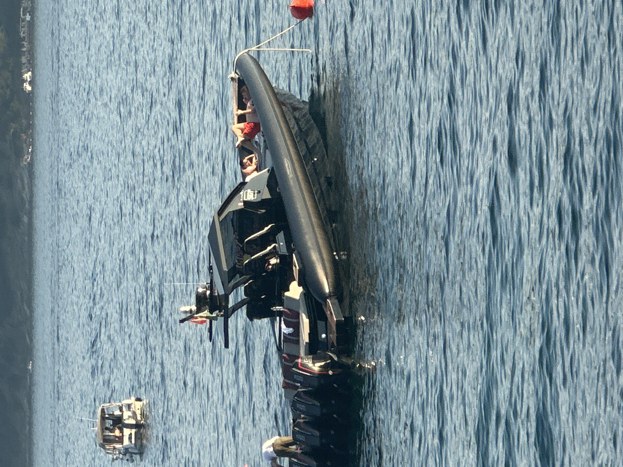 A black rigid inflatable boat (RIB) with multiple outboard engines is anchored in calm, blue water. Three individuals are visible on the boat. One person, wearing a white shirt and brown pants, is standing near the stern of the boat, appearing to be attending to the engines. Two other individuals are lying down on the forward deck of the RIB, one of whom is wearing red shorts. A red buoy is tethered to the boat by a rope and floats nearby. In the background, another smaller boat with a canopy is visible, and a distant shoreline with buildings and vegetation can be seen under a hazy sky. The scene appears to be set in the waters off Budva, Montenegro.