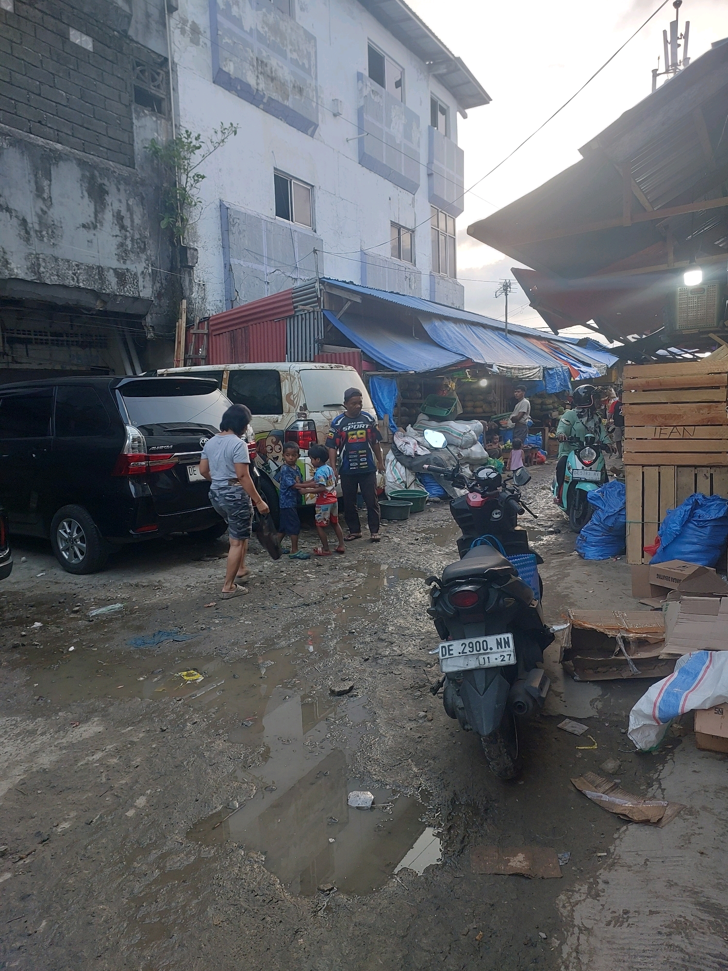 The image depicts an outdoor market scene in Ambon, Indonesia, characterized by muddy terrain and overcast skies. A black SUV is parked on the left side of the frame, with its rear facing the viewer. Beside it, a woman wearing a gray t-shirt and camouflage shorts is walking away from the camera, carrying a dark bag. Three young boys, dressed in casual clothing, are standing near the SUV, looking towards the market area.
Further into the market, several motorbikes are visible, some parked and others in use. One light blue motorbike with the license plate "DE 5783 NV" is being ridden by a person wearing a helmet and a light green jacket. Another motorbike, dark colored with the license plate "DE 2900 NN" and the inscription "01-27", is parked in the foreground.
The market stalls are covered with blue and red tarpaulins, creating shaded areas. Piles of goods, including what appear to be coconuts, are visible under the tarpaulins. Wooden crates are stacked on the right side of the image, with one crate bearing the stencil "LEAN". Several large blue bags are placed on the ground near the crates.
The ground is a mixture of mud and dirt, with several puddles of water reflecting the surroundings. Debris, including plastic bags and cardboard, is scattered throughout the area, suggesting a busy and somewhat unkempt environment. The background shows a multi-story building with a light blue facade, showing signs of wear and tear. The overall impression is that of a functional, but potentially challenging, marketplace.