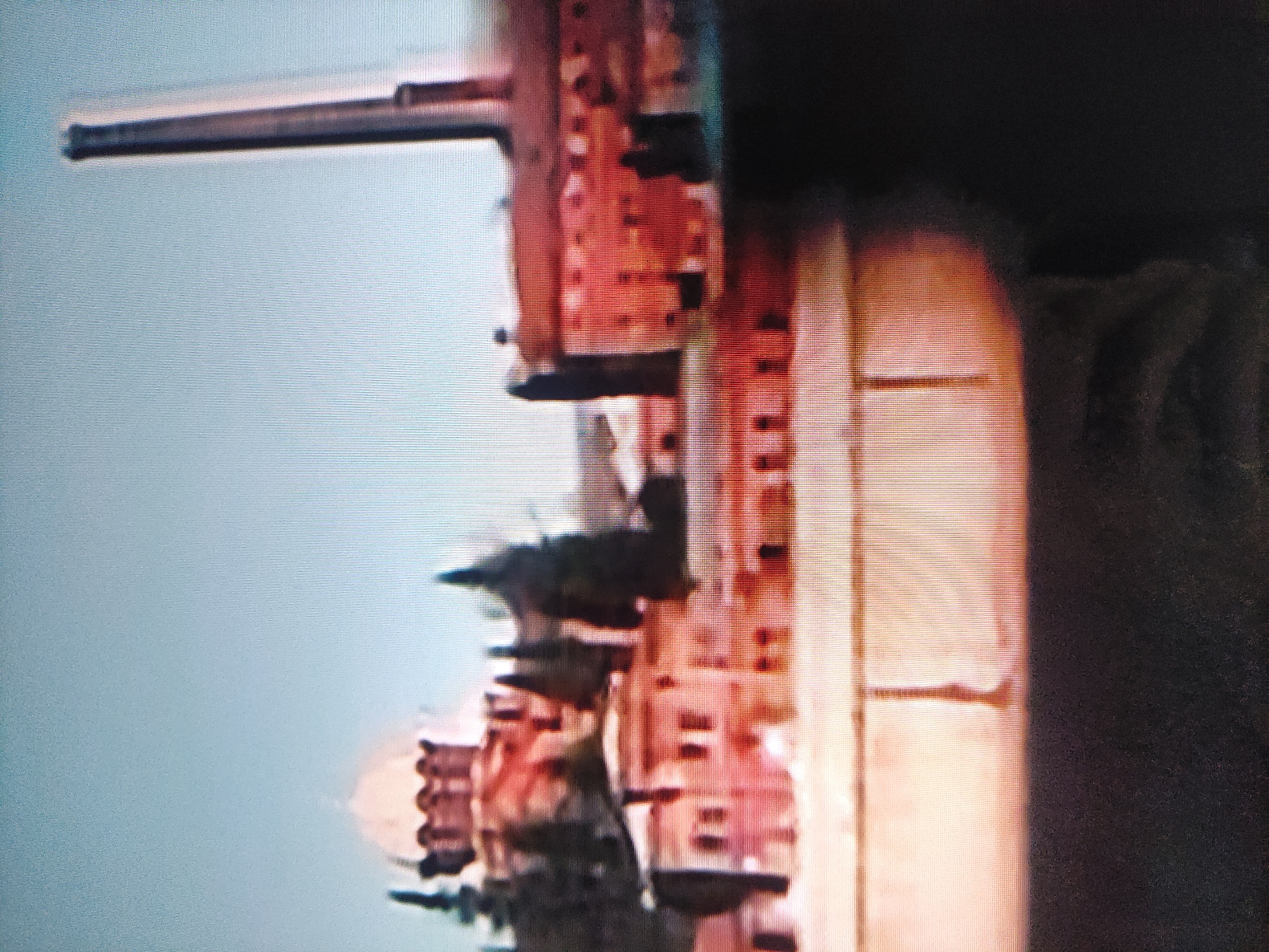 The image shows the Cathedral of Archangel Michael in Pavlodar, Kazakhstan. The cathedral is a brick building with multiple levels and towers. A long, thin object extends horizontally from the top left of the frame towards the building, possibly a light fixture. To the right, the background is obscured by darkness. The sky is visible on the left side of the building. The image appears to be taken from a distance.