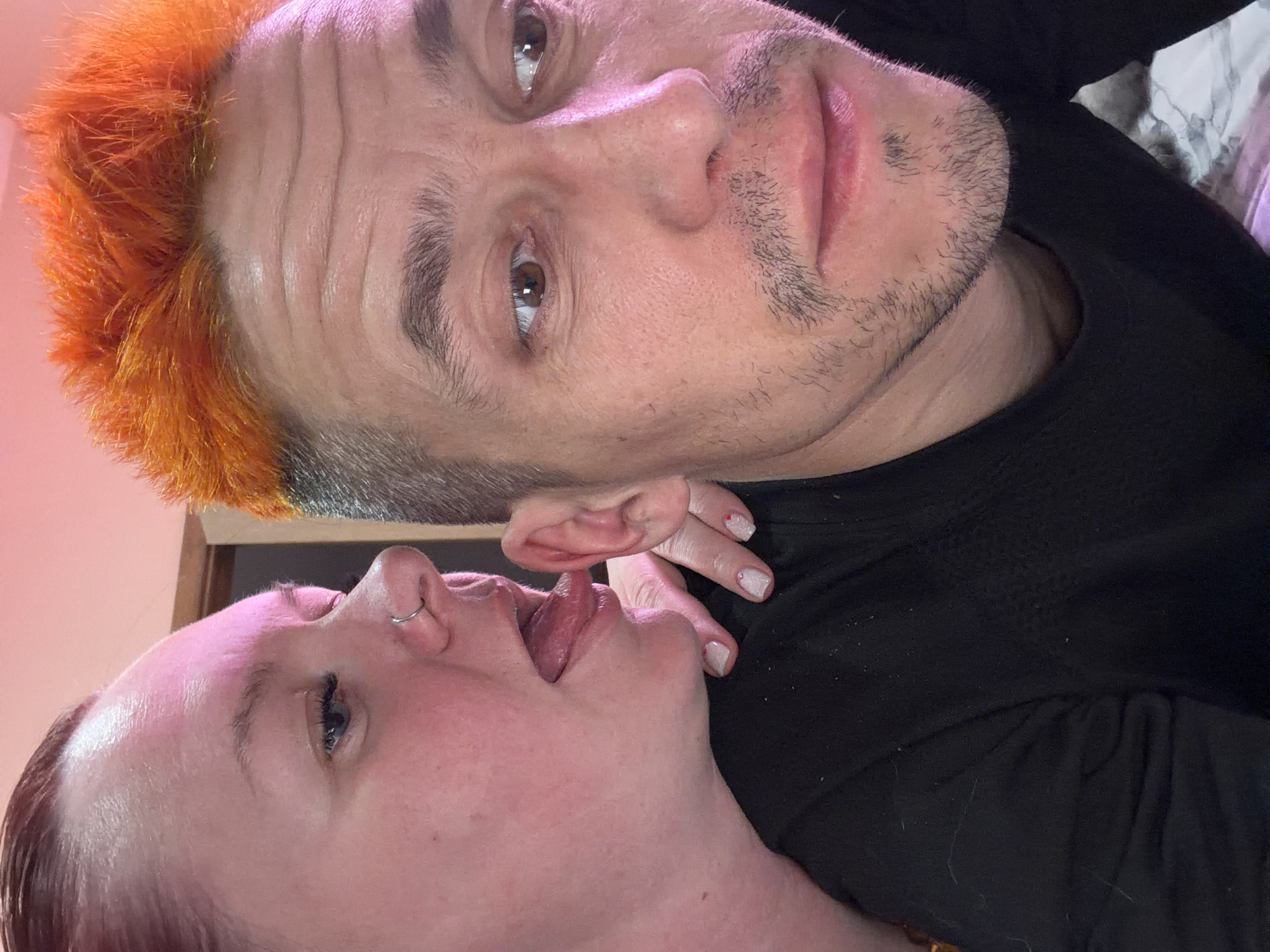 Two people are in close proximity. One has orange hair and a short haircut. The other has reddish-brown hair and is lying beneath the first person. The person underneath has their mouth open. A hand with manicured nails is visible near the lower person's ear. Both are wearing black clothing. The location is not discernible from the given image.