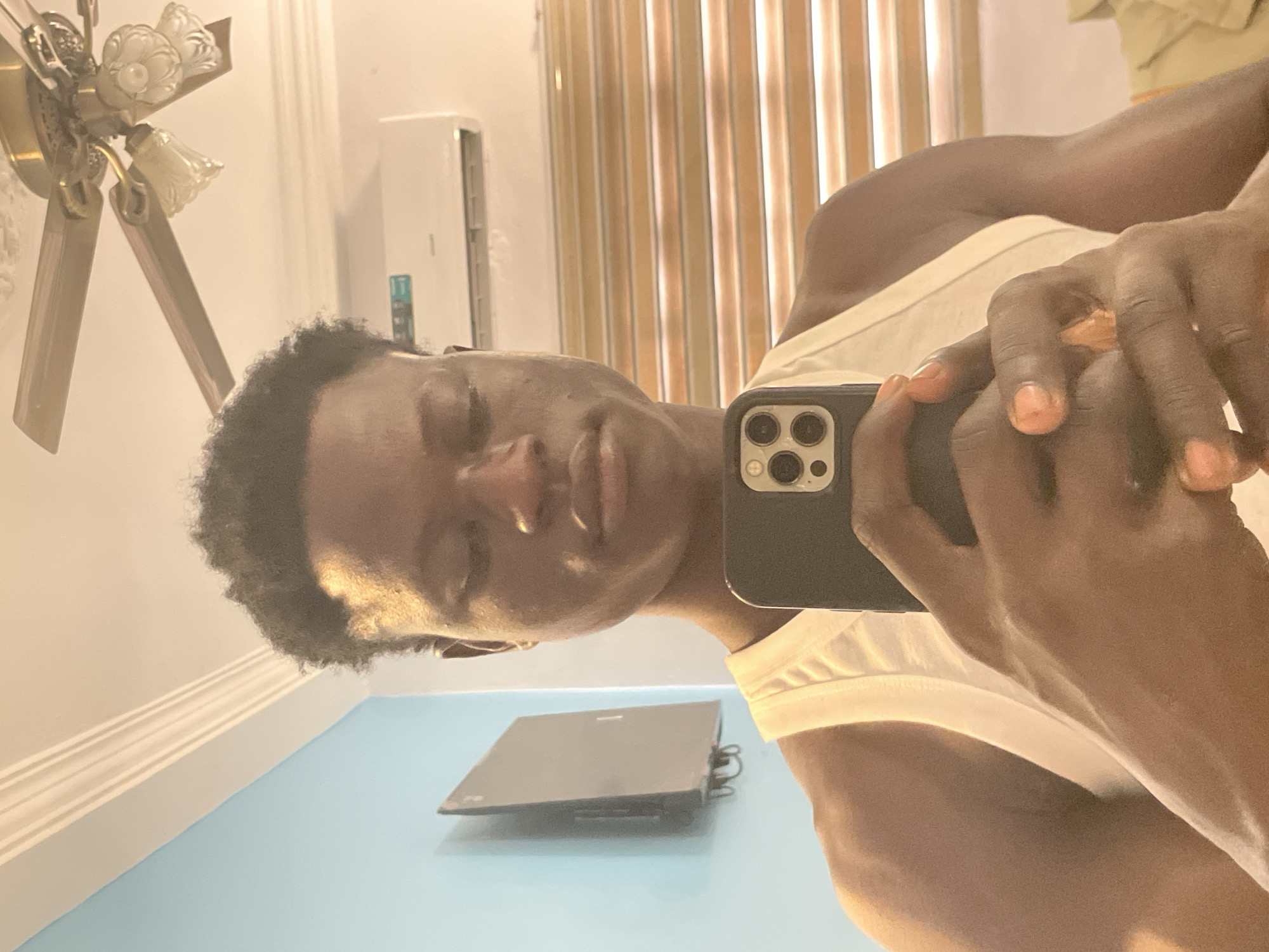 A person takes a selfie in a room. The person has dark skin, short, dark hair, and is wearing a white tank top. They are holding a smartphone in their hand. Behind the person are window blinds. An air conditioner is visible on the wall, as is a ceiling fan. The presence of the air conditioner suggests an indoor environment, and the type of ceiling fan appears common in many residences. There is a countertop reflecting light, and what appears to be an object placed on it. There are no elements in the image that specifically indicate the location in the city of Badagry, Nigeria.