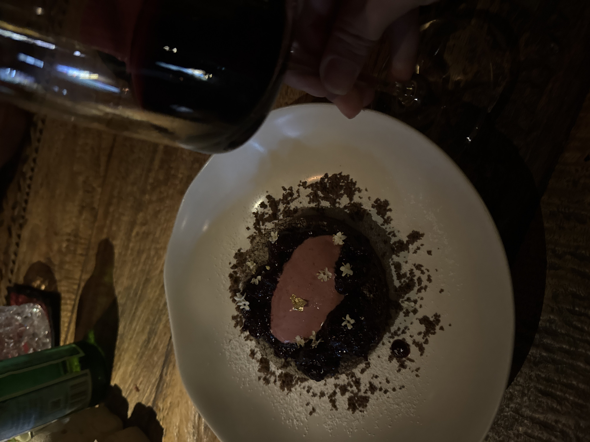The image presents a dimly lit, top-down view of a sophisticated dessert served on a white, irregularly shaped plate, set on a dark wooden table in what appears to be a restaurant or bar in North Kuta, Indonesia.
The dessert is centrally placed and features a dark, rich base, possibly a berry compote or chocolate creation. It is artfully topped with a light pink cream or mousse, highlighted by a small, shimmering piece of gold leaf. Small white blossoms, resembling elderflowers, are carefully arranged on and around the pink topping, adding an elegant garnish. The plate is adorned with dark, crumbly sprinkles, likely chocolate or cookie crumbs, and a delicate dusting of powdered sugar around the edges.
In the upper left portion of the image, a hand holds a dark glass bottle or wine glass, positioned over the plate, suggesting the action of pouring a dark liquid, possibly wine or a sauce, or simply being held nearby. Reflections on the glass indicate ambient light sources in the room.
To the bottom left, parts of a green glass bottle and another reflective object are visible, further hinting at a dining or bar setting. The overall ambiance is intimate and focused on the culinary presentation, indicative of an evening meal or dessert experience. There is no visible text in the image.
