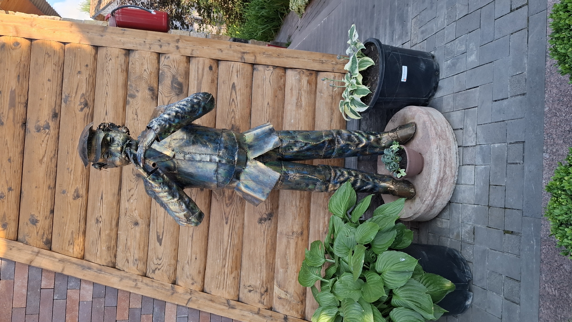 This is a medium shot of a metal sculpture of a man, presented in a vertical orientation. The sculpture, made of dark metal with iridescent blue and green highlights, depicts a man in a military-style uniform with a cap. He is posed with his hands at his sides, seemingly walking or standing at attention.
The sculpture is placed on a round, beige, stone-like base, which itself rests on a dark grey brick pathway. To the left of the sculpture is a rustic wooden wall, constructed from vertical planks. At the top of the wooden wall, a red object, possibly a fuel canister or a toolbox, is visible.
In the background, to the right of the sculpture, a large black planter holds a variegated hosta plant. Another smaller, brown pot with a small succulent is placed on the stone base, between the sculpture's feet. In the foreground, to the right and bottom of the frame, more lush green hosta plants are visible, along with a hint of a dark plastic pot. The scene is set outdoors, with a mix of paving stones, wooden structures, and greenery suggesting a garden or an outdoor decorative area.
The lighting indicates it is daytime, likely sunny, casting subtle shadows. The overall mood is one of outdoor display and decoration. There is no discernible text in the image, apart from a small white label on the large black planter, which is too blurry to read. The location context provided is Gyulagarak, Armenia.