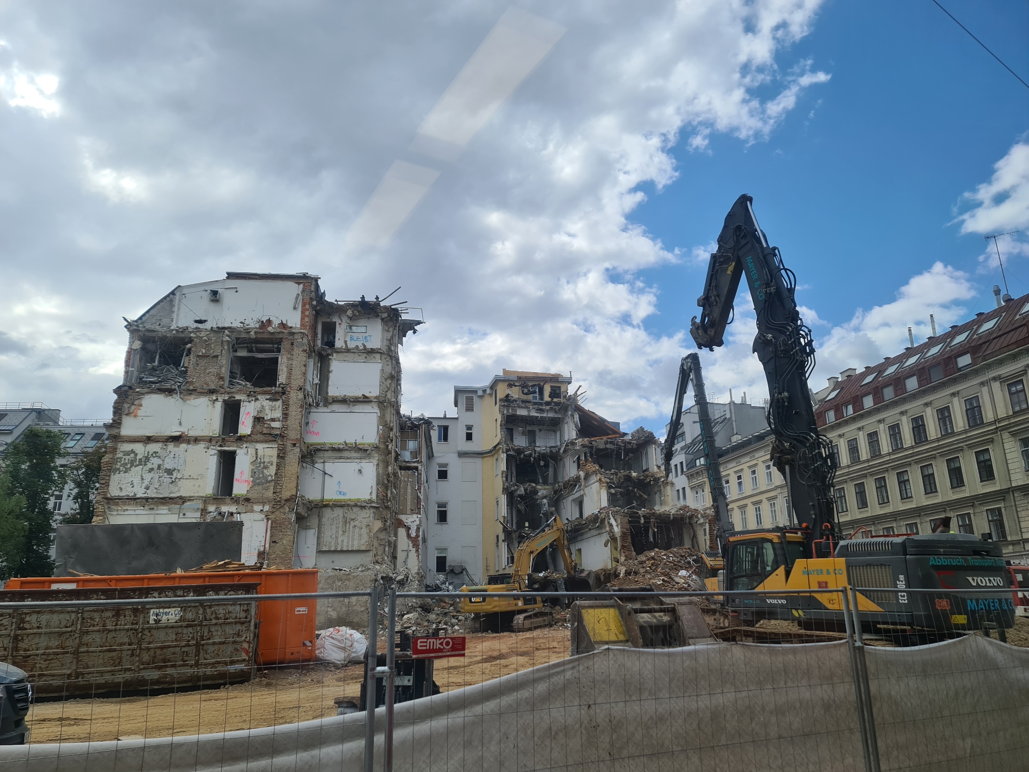 This media file depicts a demolition site in Vienna, Austria. The primary subjects are the partially demolished multi-story buildings, surrounded by construction debris and heavy machinery. Two excavators are prominently visible, one a large black demolition excavator with the branding "MAYER & CO" and "VOLVO" and the other a yellow tracked excavator, likely a CAT. Large orange and grey dumpster bins are present, along with piles of rubble. A construction fence with netting encloses the area, with a white tarp-covered object visible behind it.
The scene suggests an active demolition or deconstruction process is underway. The sky is partly cloudy with patches of blue, indicating daytime. There are no people clearly visible in the foreground or engaged in any activity.
Notable details include the visible text on the machinery: "MAYER & CO", "VOLVO", and "Abbruch, Transport, Rec" (likely German for "Demolition, Transport, Recycling"). There are also various markings on the buildings themselves, such as "CEVI" and "BLEIBT". The architectural style of the surrounding buildings in the background, particularly the one on the right with its dormer windows and mansard roof, is characteristic of older European cities, consistent with the Vienna location. The overall impression is one of industrial activity and urban renewal.