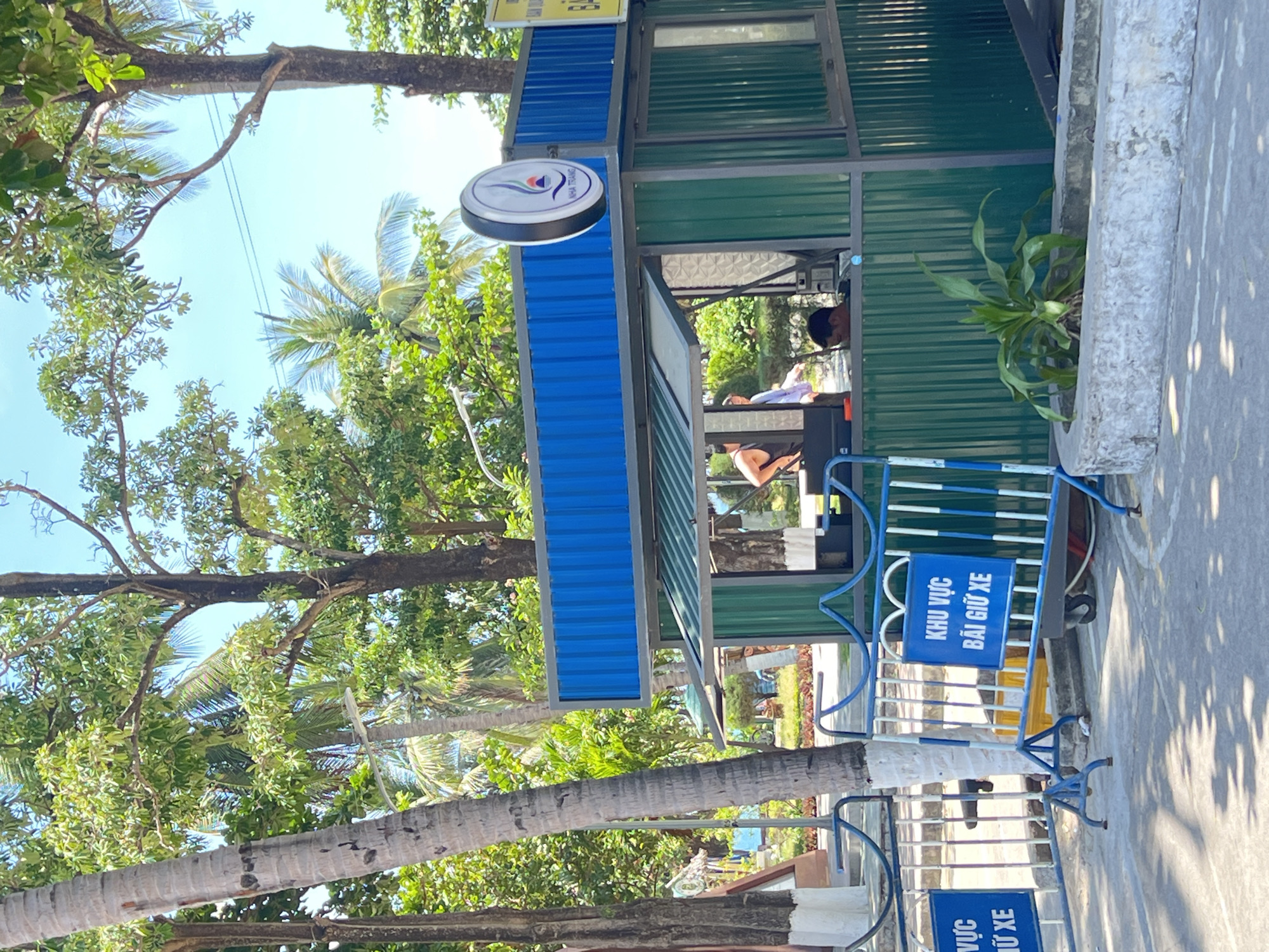 The image shows a blue and green corrugated metal booth next to a paved walkway in Nha Trang Ward, Vietnam. The booth has a circular sign with a white and blue logo on top. There is a blue metal barricade in front of the booth with a sign in Vietnamese reading "KHU VUC BAI GIU XE" (Parking Area). Two people are visible inside the booth. The environment consists of tropical trees and foliage, indicating a coastal area.
