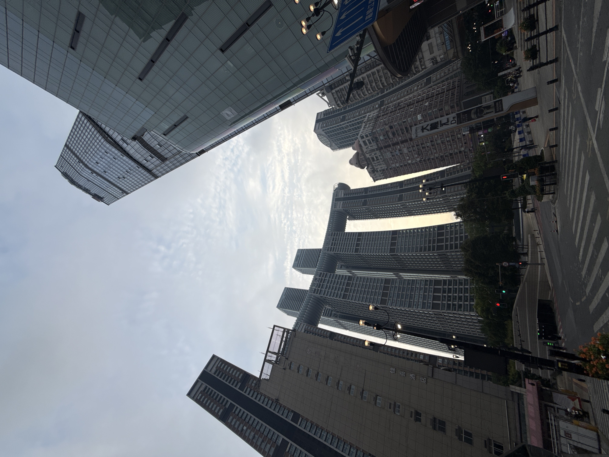 The image depicts a city scene with a unique architectural landmark. Dominating the frame are several exceptionally tall skyscrapers, angled towards the sky. The most distinctive structure is a cluster of four interconnected towers, with a horizontal bridge-like element spanning between the tops of two of them, creating a distinctive gateway appearance. The skyscrapers are characterized by their glass facades and rectilinear designs. In the foreground, a street is visible with pedestrian crossings and traffic lights. A street lamp with multiple lights stands prominently on the left side of the frame. The sky above is overcast with scattered clouds. The architecture suggests a modern urban environment, likely a commercial or residential district in a major city. The presence of a street lamp and traffic signals indicates a functional public space. The overall impression is of a contemporary cityscape with an emphasis on verticality and bold architectural design. The location is Yuzhong District, China.