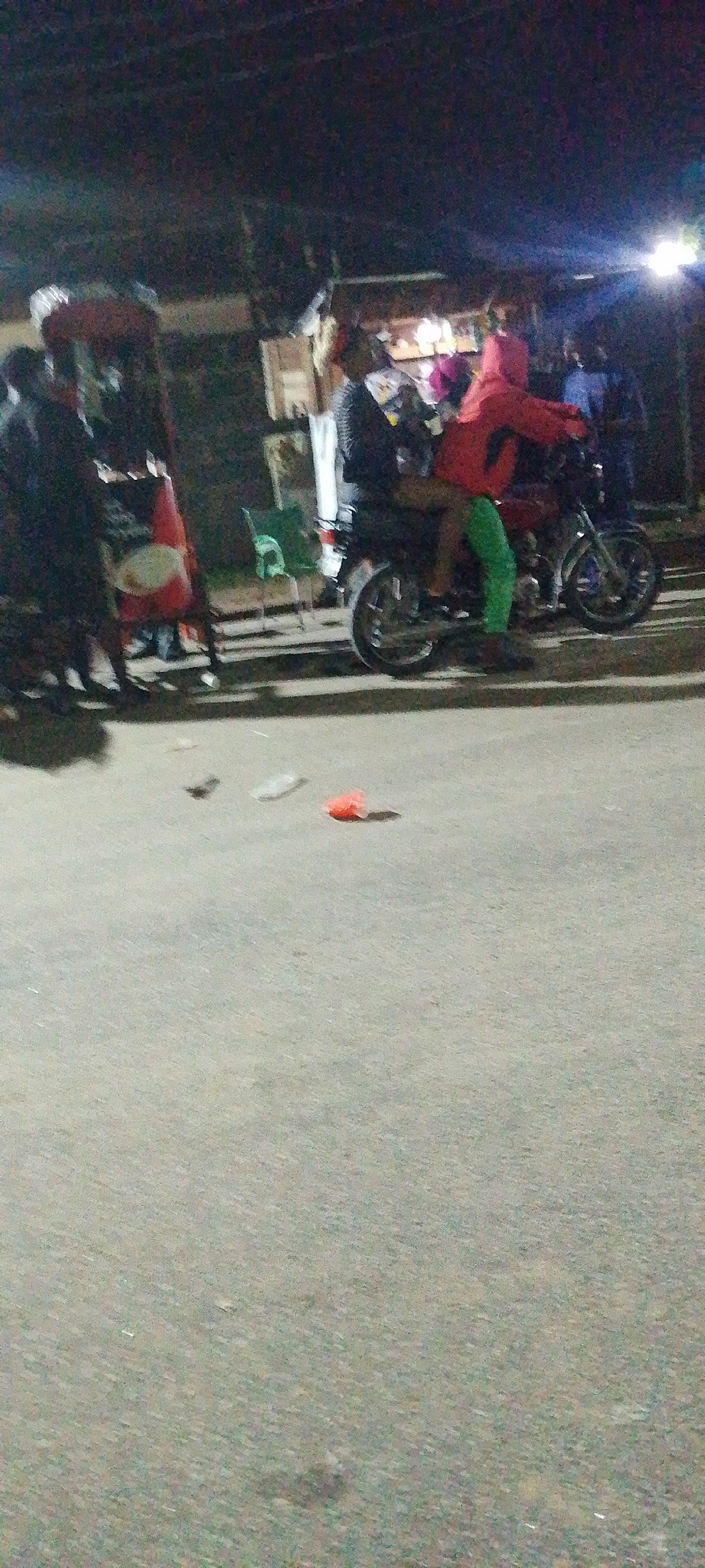 The image depicts a nighttime scene in Lokoja, Nigeria. Two individuals are on a motorcycle. The driver wears a dark jacket, while the passenger is clad in a red hoodie and green pants. A group of people stand near a makeshift stall or shop. There are a few chairs visible as well. The foreground shows a paved road with litter. The overall illumination is poor, with light sources concentrated around the shop area.