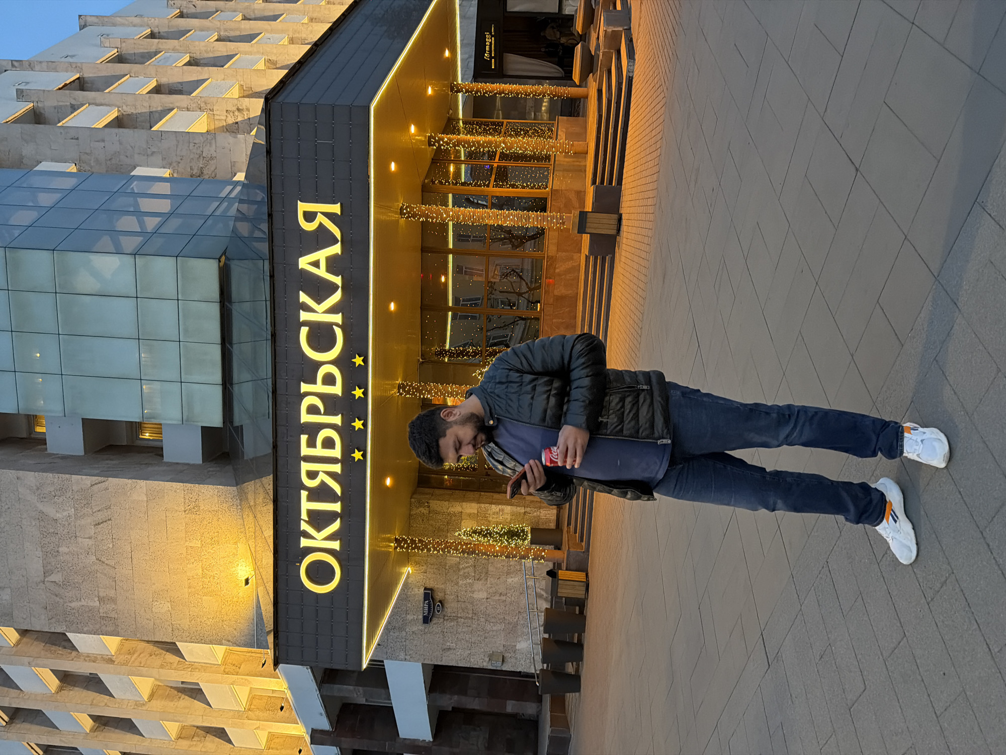 A man stands on a sidewalk in front of the Oktyabrskaya Hotel, located in Krasnoyarsk, Russia. He is holding a cell phone in his left hand and a can of Coca-Cola in his right. The hotel entrance is decorated with golden light strands, and a sign displaying the hotel's name in Cyrillic script and with golden stars is prominently visible. The sidewalk is paved with large rectangular tiles.