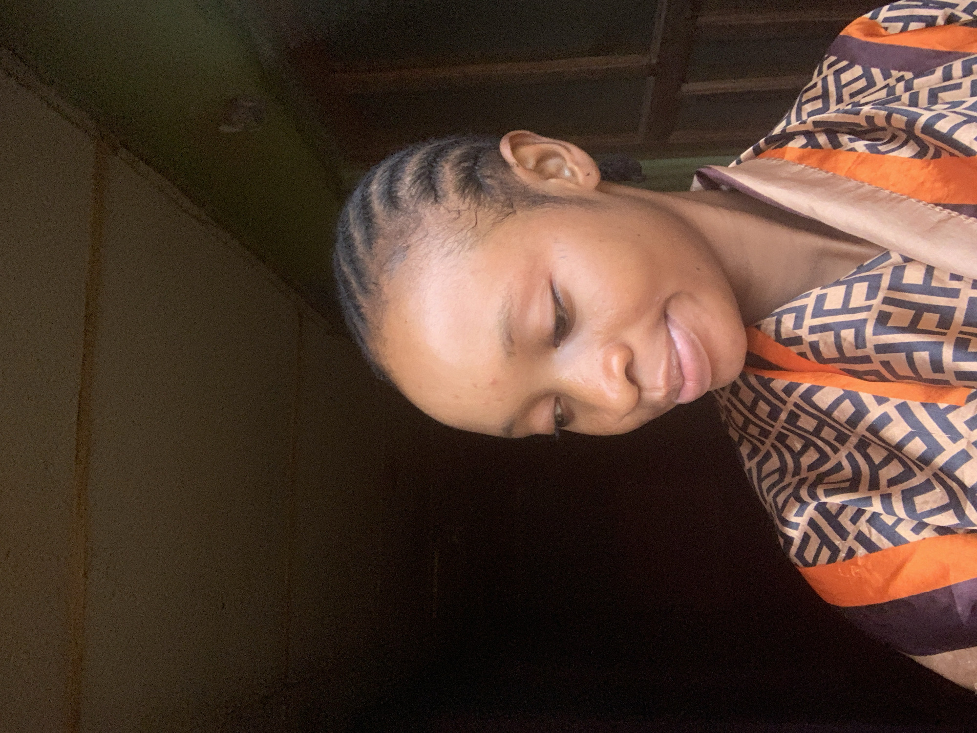 The media file features a close-up, portrait-style shot of a young Black woman with her eyes closed, appearing to be resting or in a moment of reflection. She has her hair styled in neat cornrows that frame her forehead. Her head is tilted downwards and to her right, with her face partially obscured by a patterned fabric draped around her. The fabric is a blend of warm beige and brown with an intricate geometric design in dark blue, accented by bright orange stripes along the edge.
The lighting suggests an indoor setting, possibly with natural light coming from a window or an opening to the side, casting shadows that create a sense of depth and intimacy. The background is dark and appears to be a wall, with some texture visible, possibly due to an aged or rustic finish. There are also hints of wooden structures above, suggesting a ceiling or overhead beams.
The woman's expression is serene, with a slight smile playing on her lips, conveying a feeling of peace or contentment. The overall mood of the image is calm and personal. There is no visible text in the media file. The location context of Shagamu, Nigeria, might imply a residential setting within the region. The time of day is not explicitly indicated, but the lighting does not suggest harsh midday sun, leaning more towards softer, ambient light.