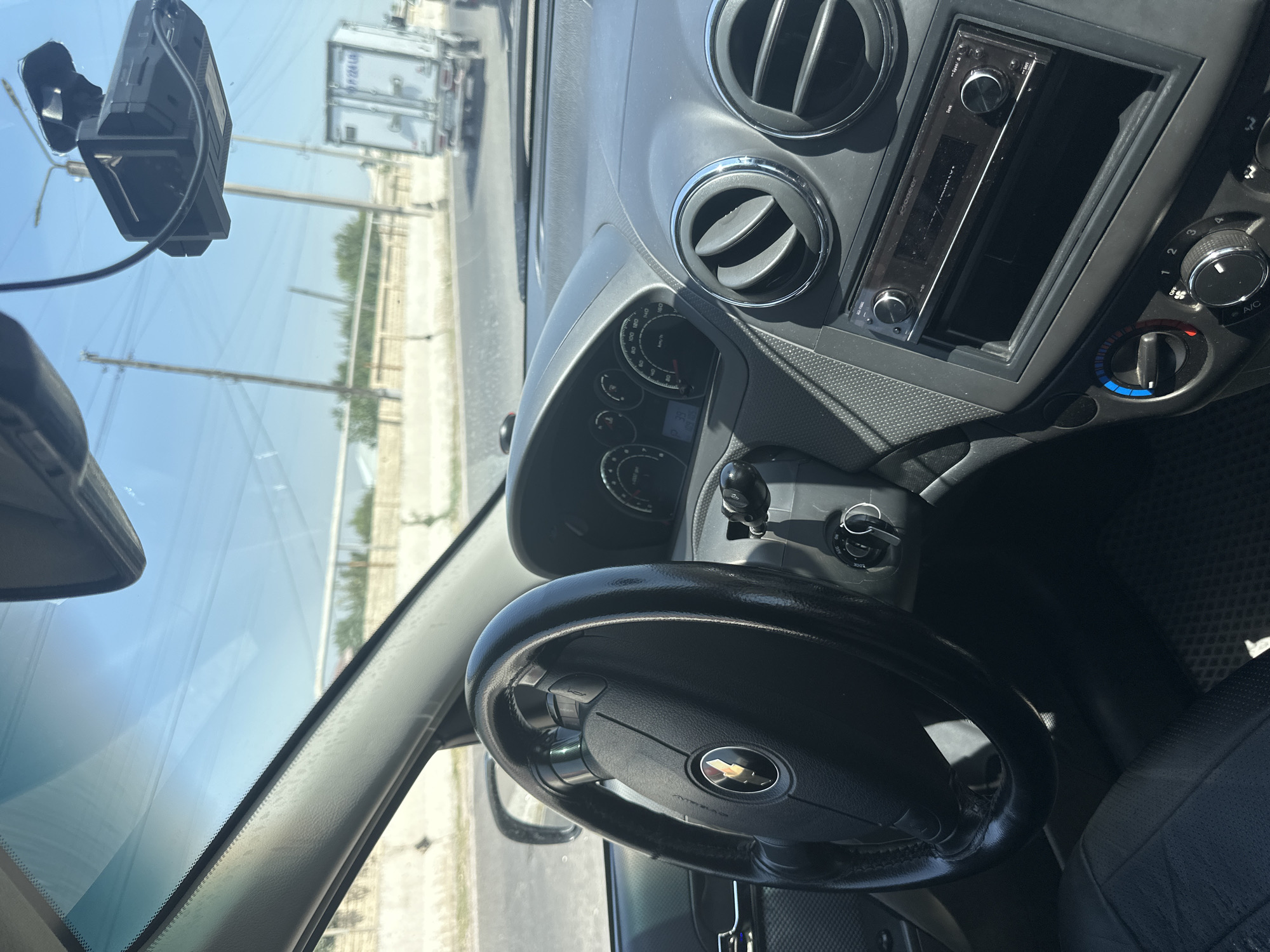 The image shows the interior of a car, likely a Chevrolet. The steering wheel with the Chevrolet logo is visible on the left, and the dashboard with the instrument cluster and air vents is on the right. A Pioneer car stereo is installed in the dashboard. Outside the car, through the windshield, a road with a truck and utility poles can be seen, suggesting an urban or suburban setting. The clear sky indicates daytime. The presence of a dashcam mounted on the windshield is also noted. Based on the context provided by the user, this scene is located in the city of Tashkent, Uzbekistan.