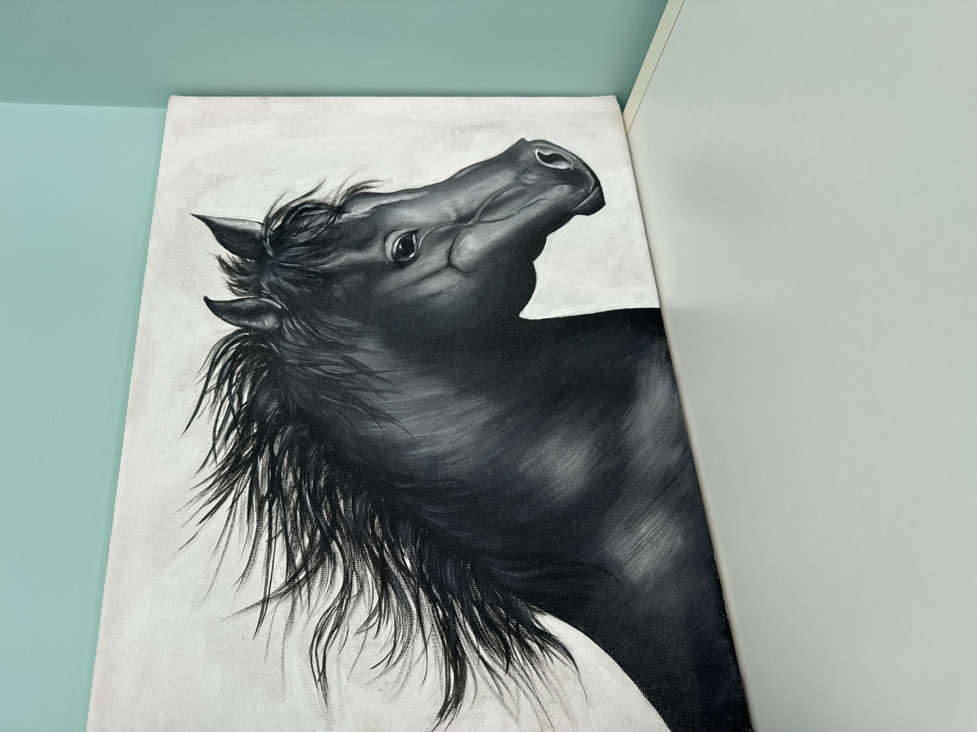 A black and white painting of a horse's head and neck, oriented diagonally, is visible in the image. The horse has a long mane, and its head is tilted upward. The painting is on a canvas, which is adjacent to a light gray wall. The background wall is a pale blue-green color. The image does not contain any information about Sofia, Bulgaria.
