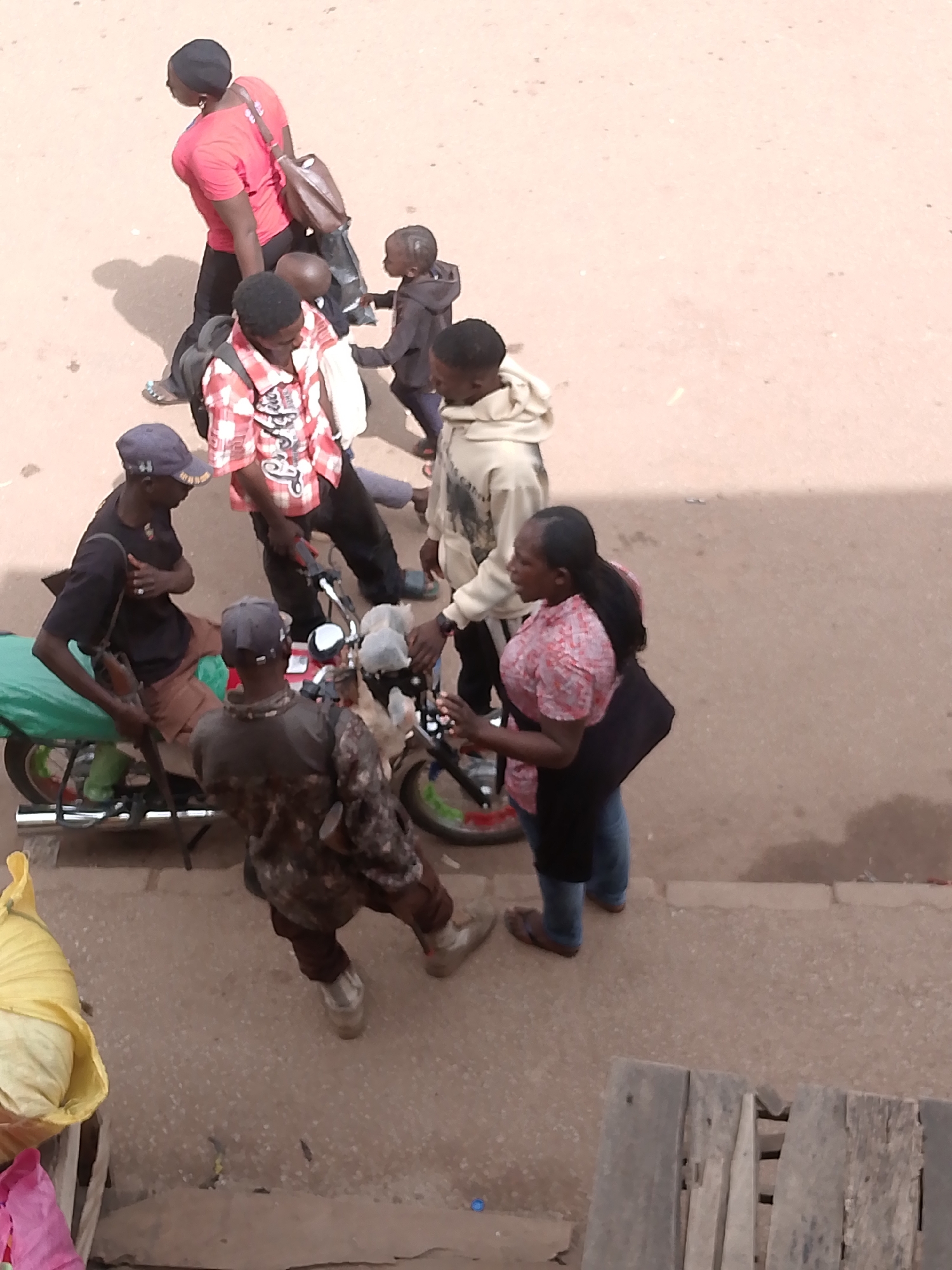The image shows a street scene in Bukuru, Nigeria. A group of people are gathered around a parked motorcycle. One man is seated on the motorcycle, while others are standing and interacting with each other. A woman wearing a patterned shirt and black skirt is facing the motorcycle, seemingly talking to someone. A man in a camouflage shirt is standing with his back to the viewer. A yellow bag is on the left side of the scene. Other individuals are seen walking away in the background. The ground appears to be unpaved, and there are some wooden planks in the foreground.